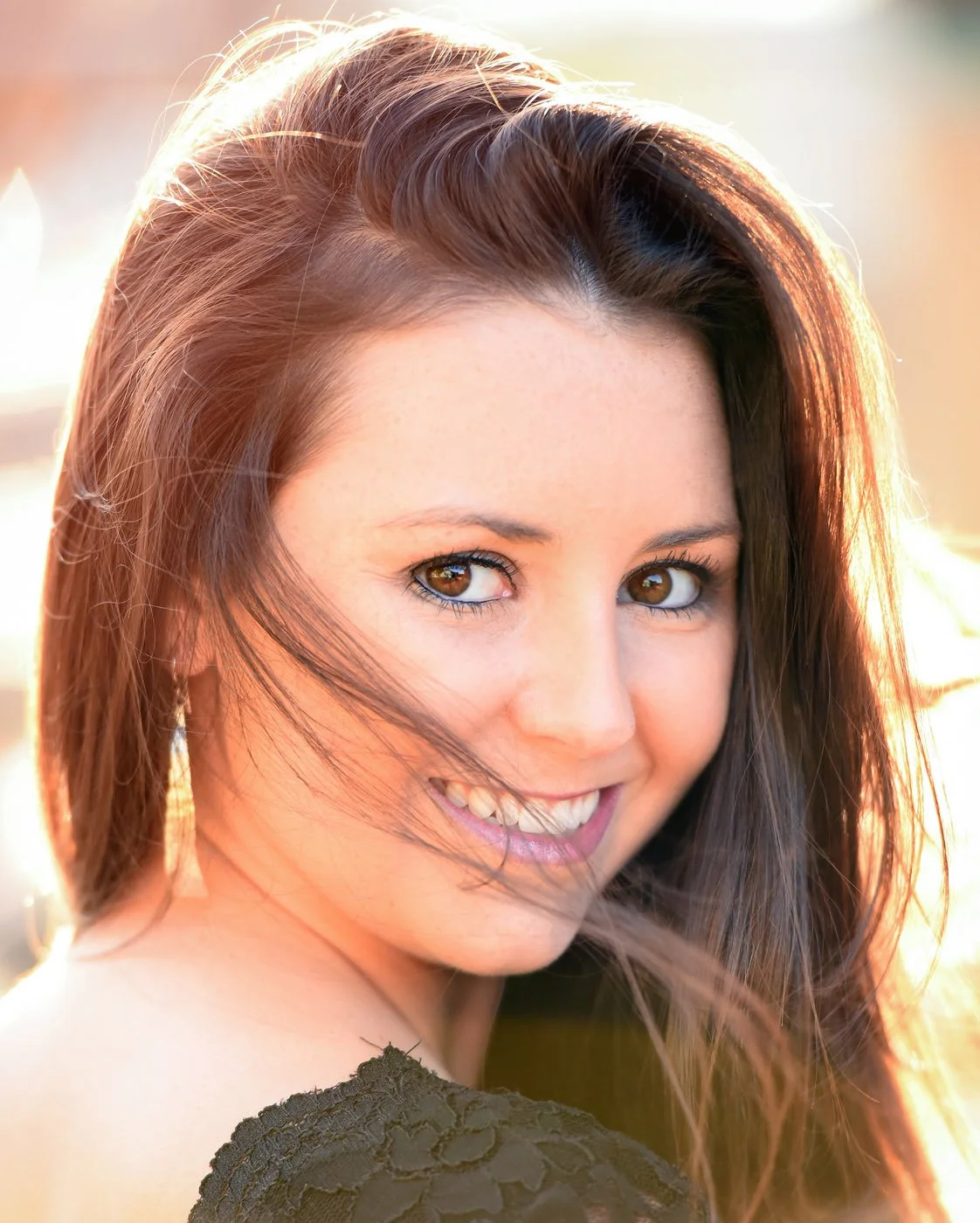 Close-up of a young woman with brown hair and brown eyes smiling outdoors at sunset.
