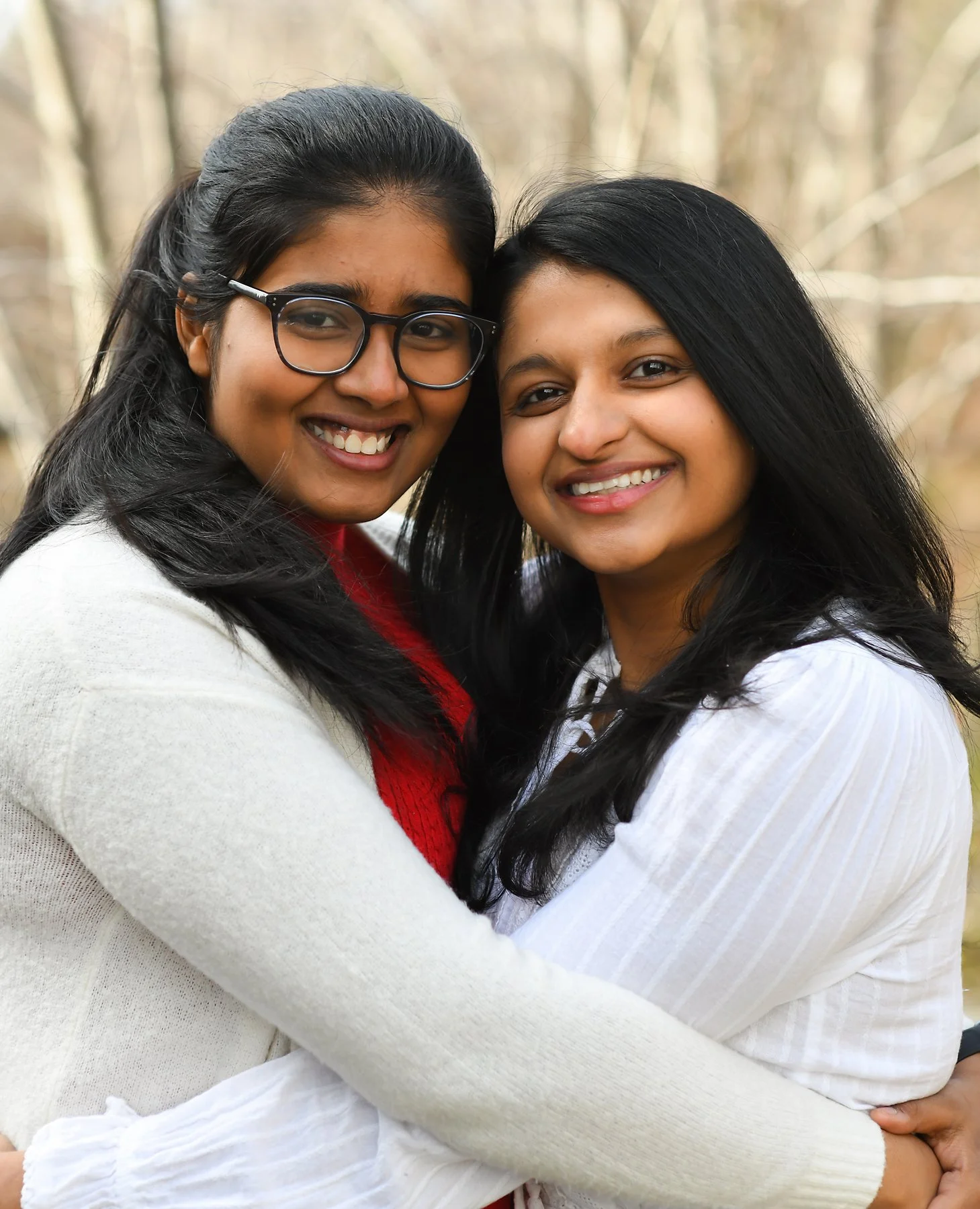 Two women smiling and hugging outdoors in a wooded area.