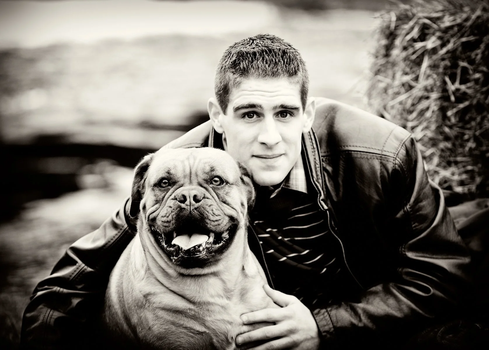 A young man with short hair and a striped shirt posing outdoors with a happy, muscular dog with a broad face, possibly a pit bull, next to him. The photo is in black and white.