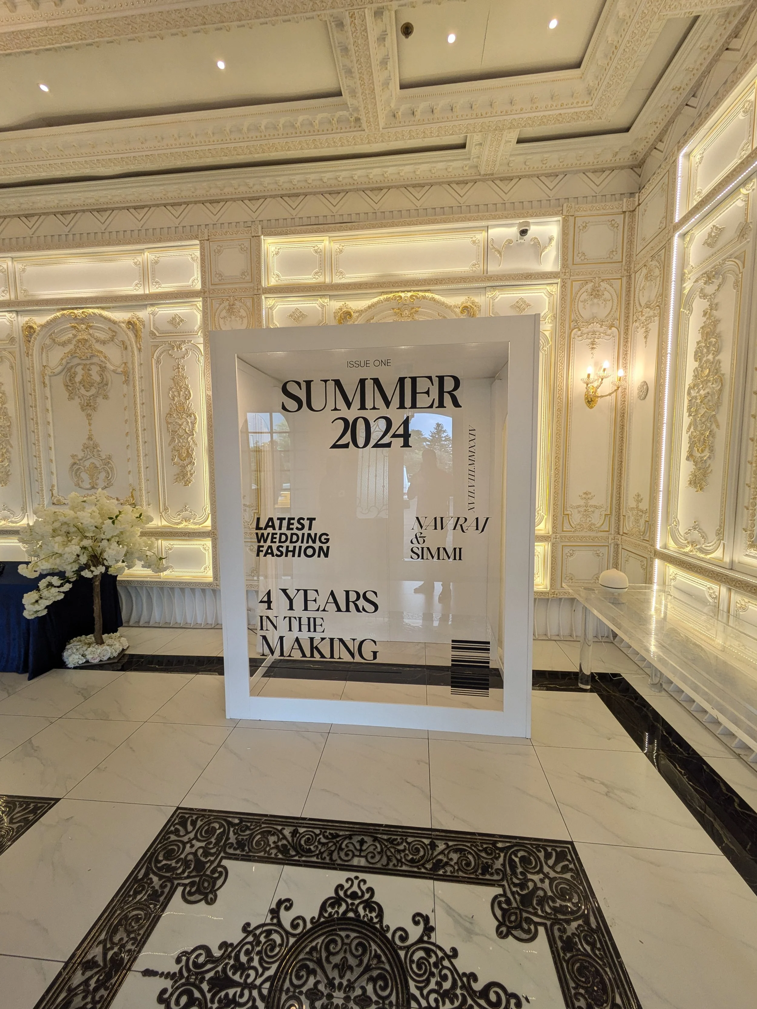 Elegant room with ornate white and gold decor, featuring a large magazine-style display case labeled "SUMMER 2024" with text about wedding fashion and a floral arrangement on the side.
