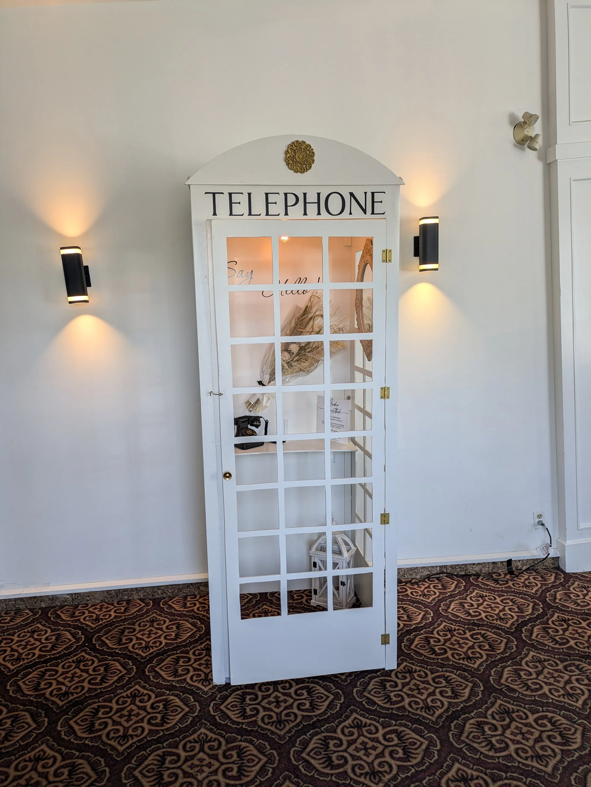 A vintage white wooden telephone booth with a glass-paneled door, labeled 'TELEPHONE' at the top, against a white wall with wall sconces and patterned carpet on the floor.