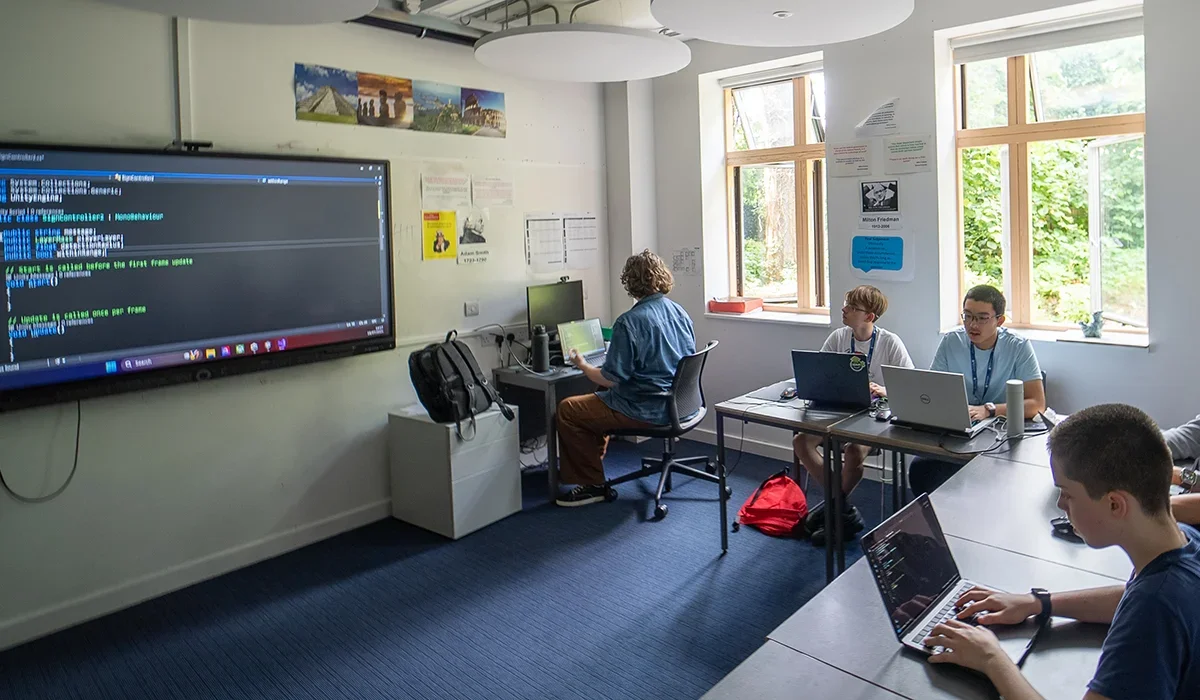 A classroom with students working on laptops, a large screen on the wall displaying code, and open windows letting in natural sunlight.