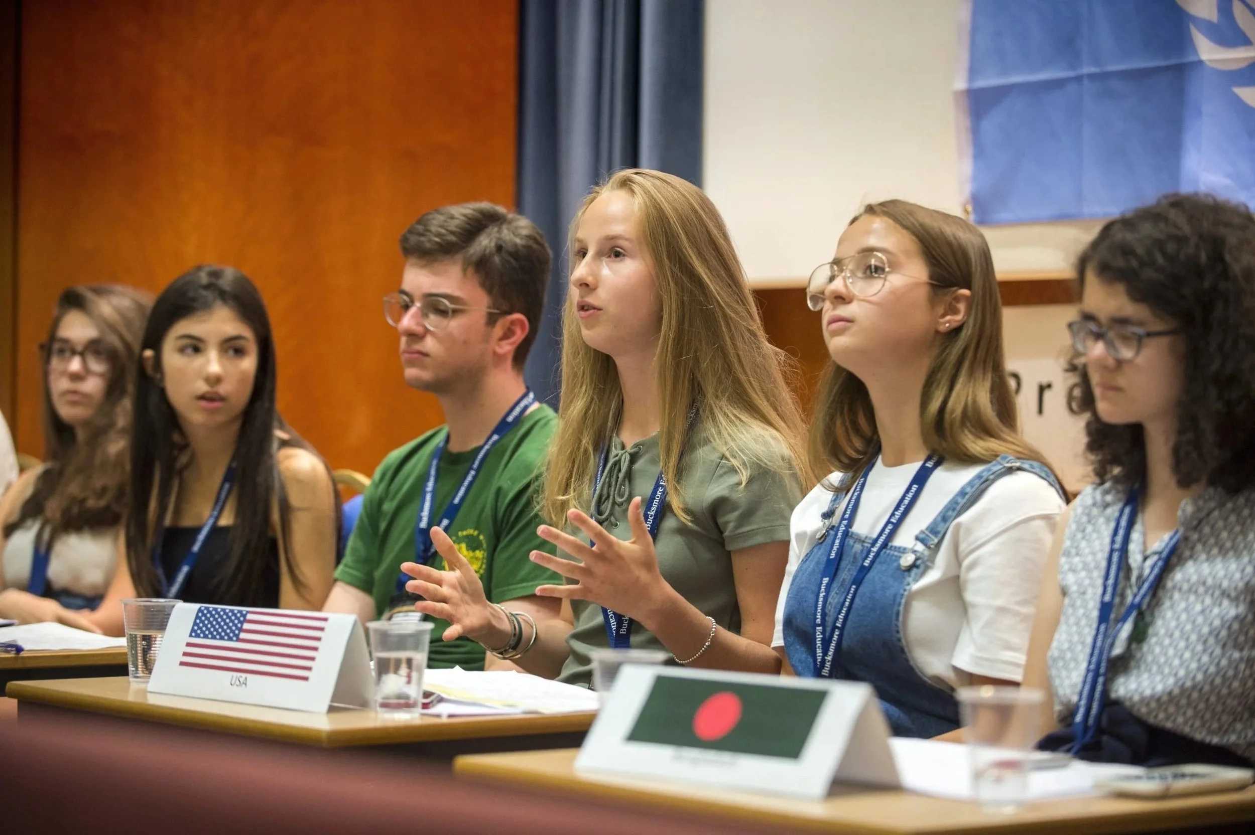 Young people sitting at a panel or conference with nameplates for USA and Bangladesh, listening and engaging in discussion.