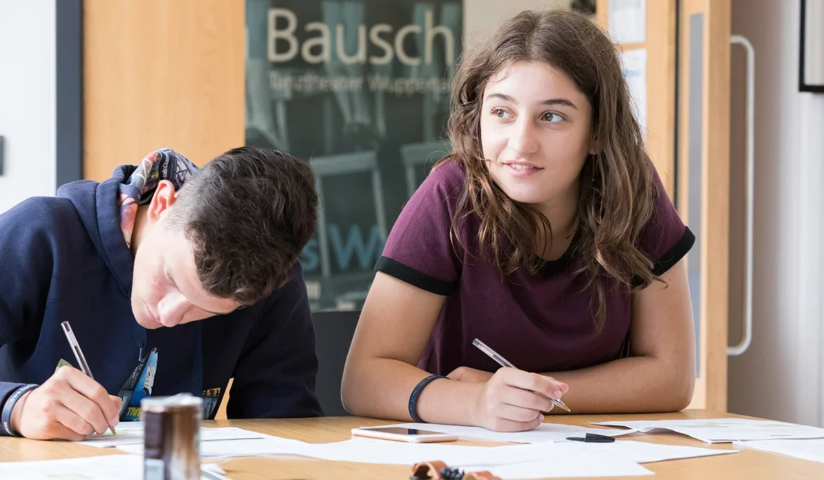 Two teenagers, a boy and a girl, sitting at a table with papers and pens, engaging in a activity in a classroom or library setting.