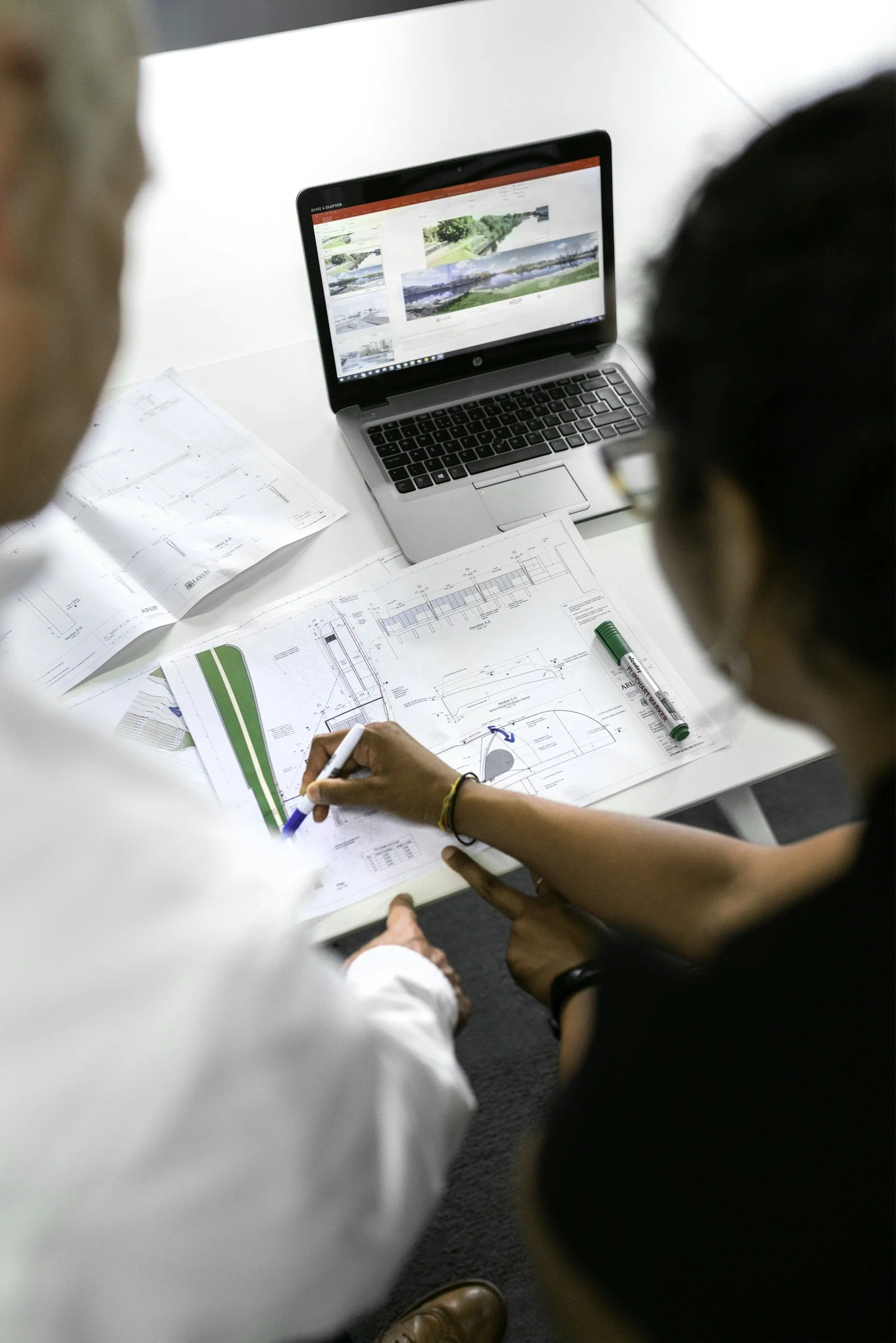 Two people are working on architectural blueprints at a white table. One is holding a pen and pointing to the blueprints while a laptop showing landscape images is open nearby.