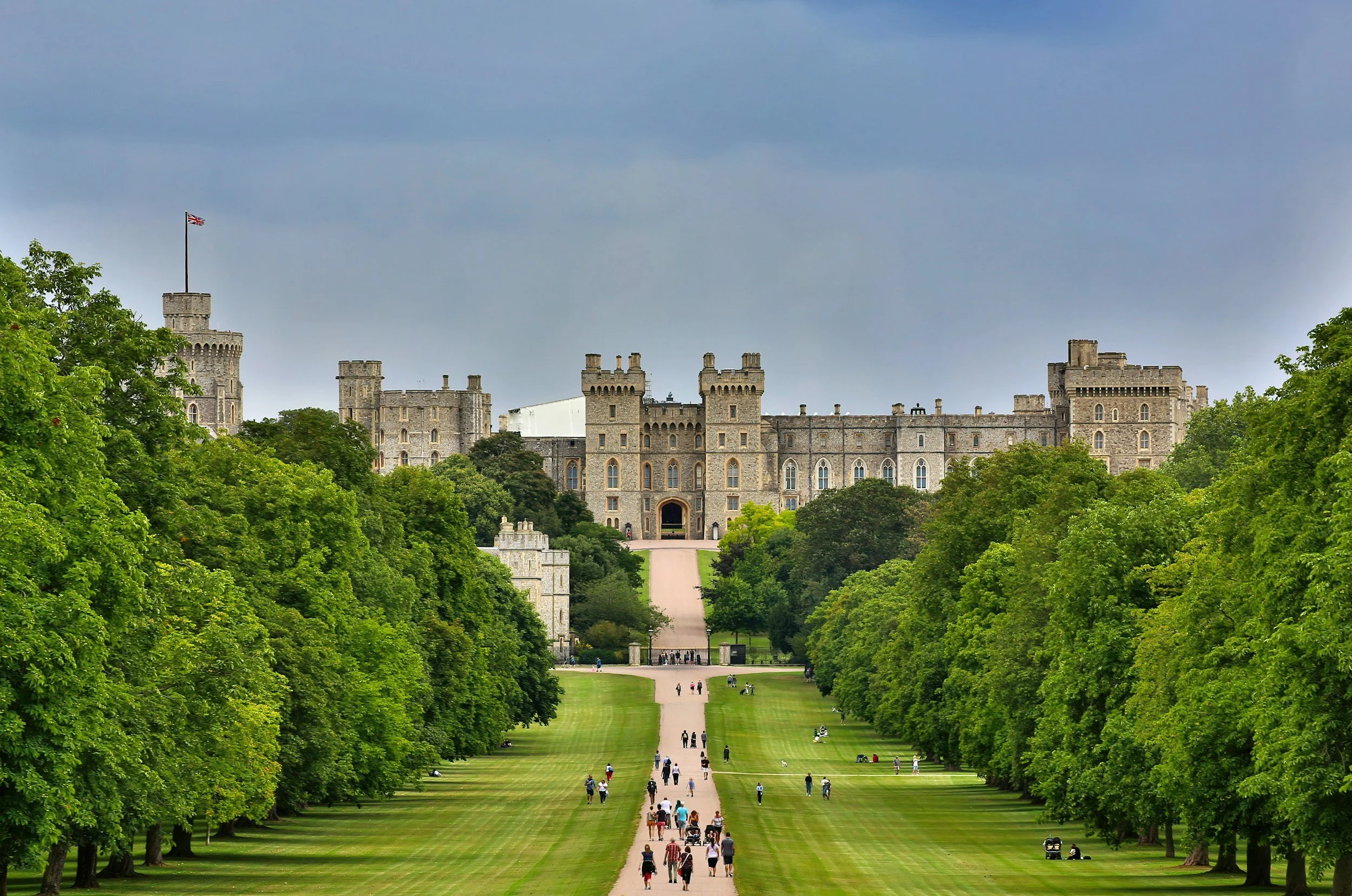 A large stone castle with multiple towers and battlements, situated atop a hill, surrounded by lush green trees, with visitors walking along a wide pathway leading up to it.
