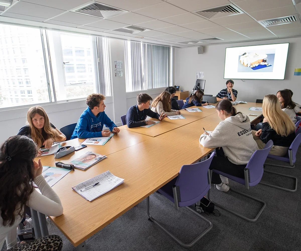 A group of students attending a class or workshop in a conference room. They are seated around a long wooden table, facing a large screen at the front of the room. The room has large windows with vertical blinds, letting in natural light.