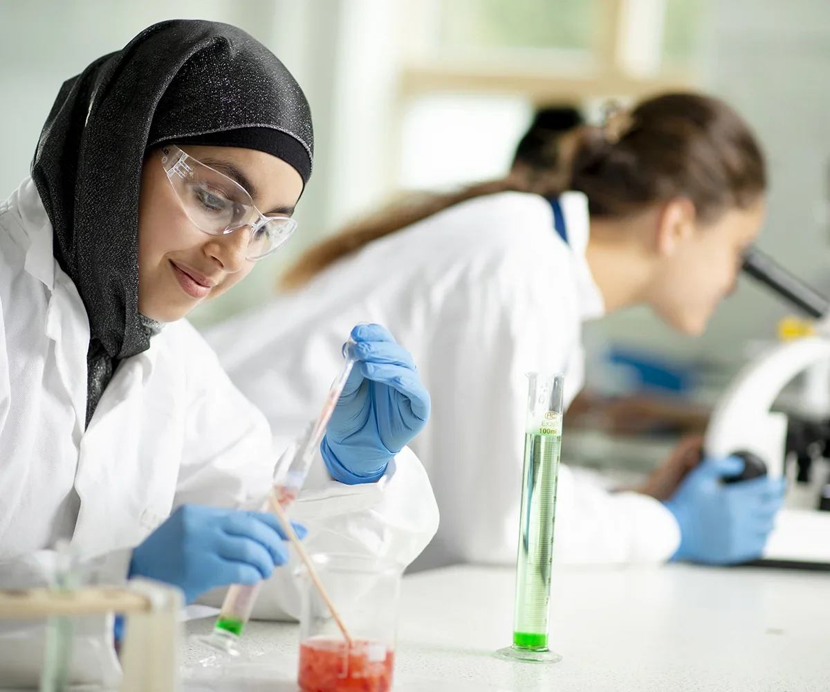 Two female scientists working in a laboratory, one wearing a black hijab, protective goggles, and blue gloves, conducting experiments with test tubes, while the other, with brown hair, looks into a microscope.