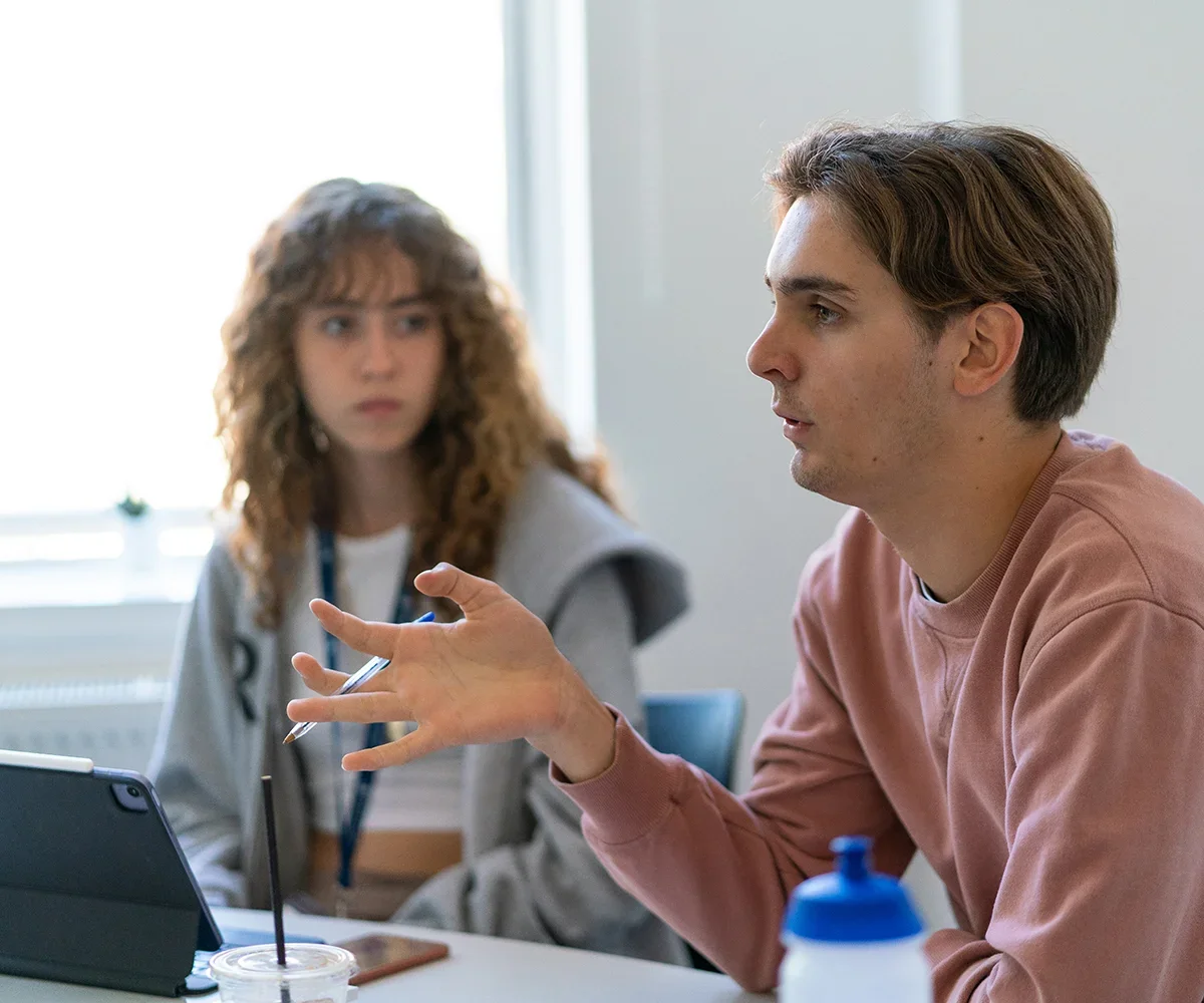 Two people sitting at a table engaged in a discussion, with one person speaking and the other listening.