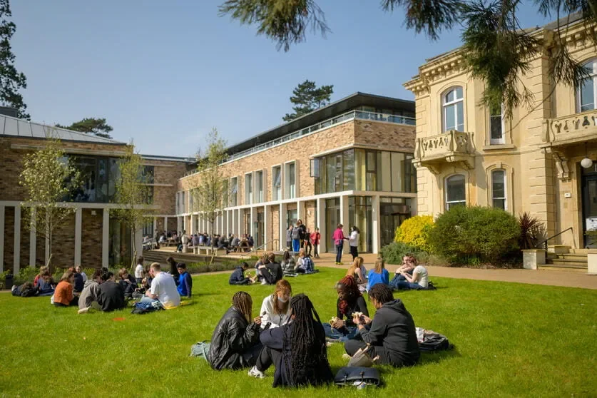 Students sitting and chatting on the grassy lawn outside a school or university building on a sunny day.