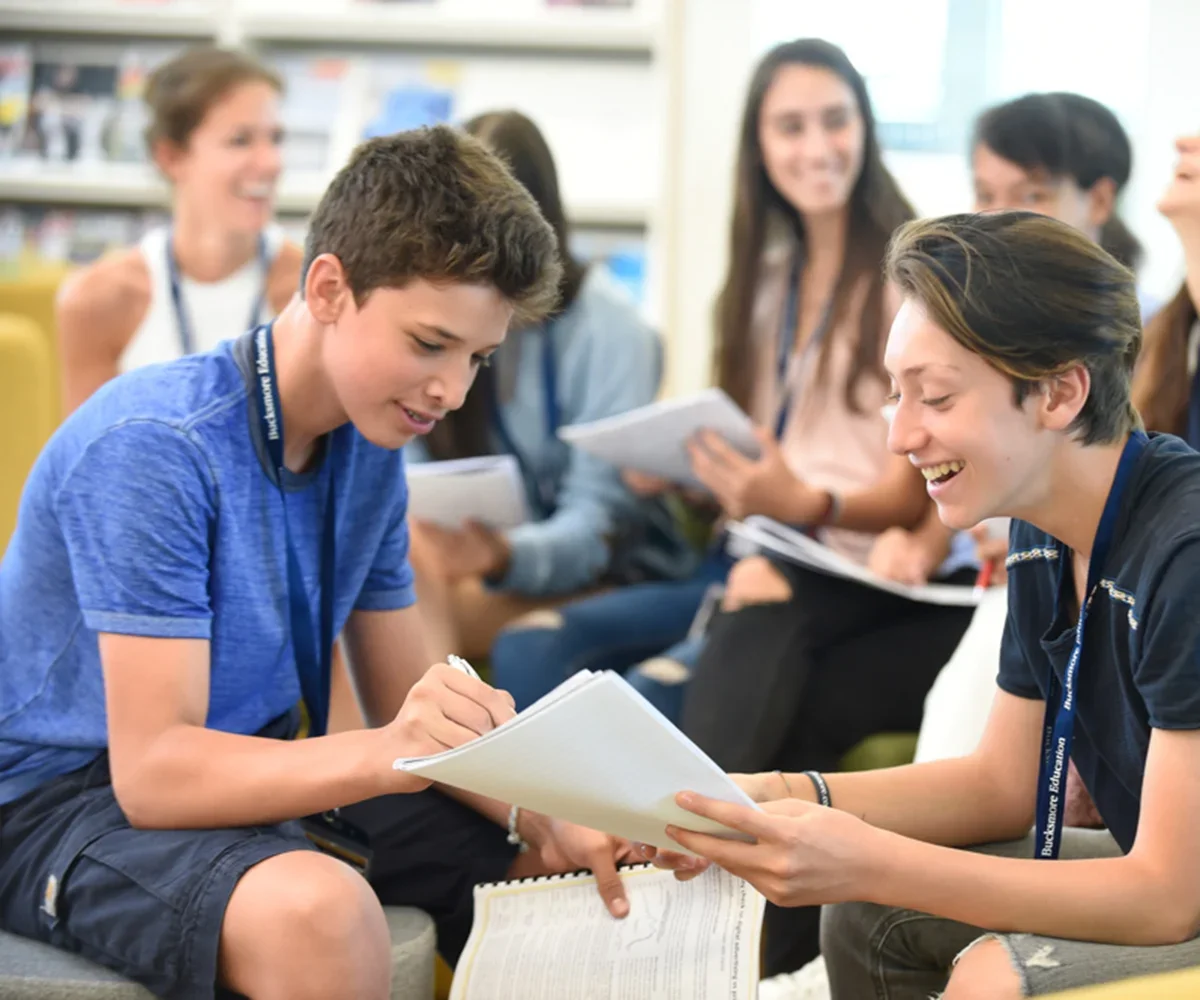 Group of smiling teenagers sitting in a circle during a discussion or activity in a classroom or library.