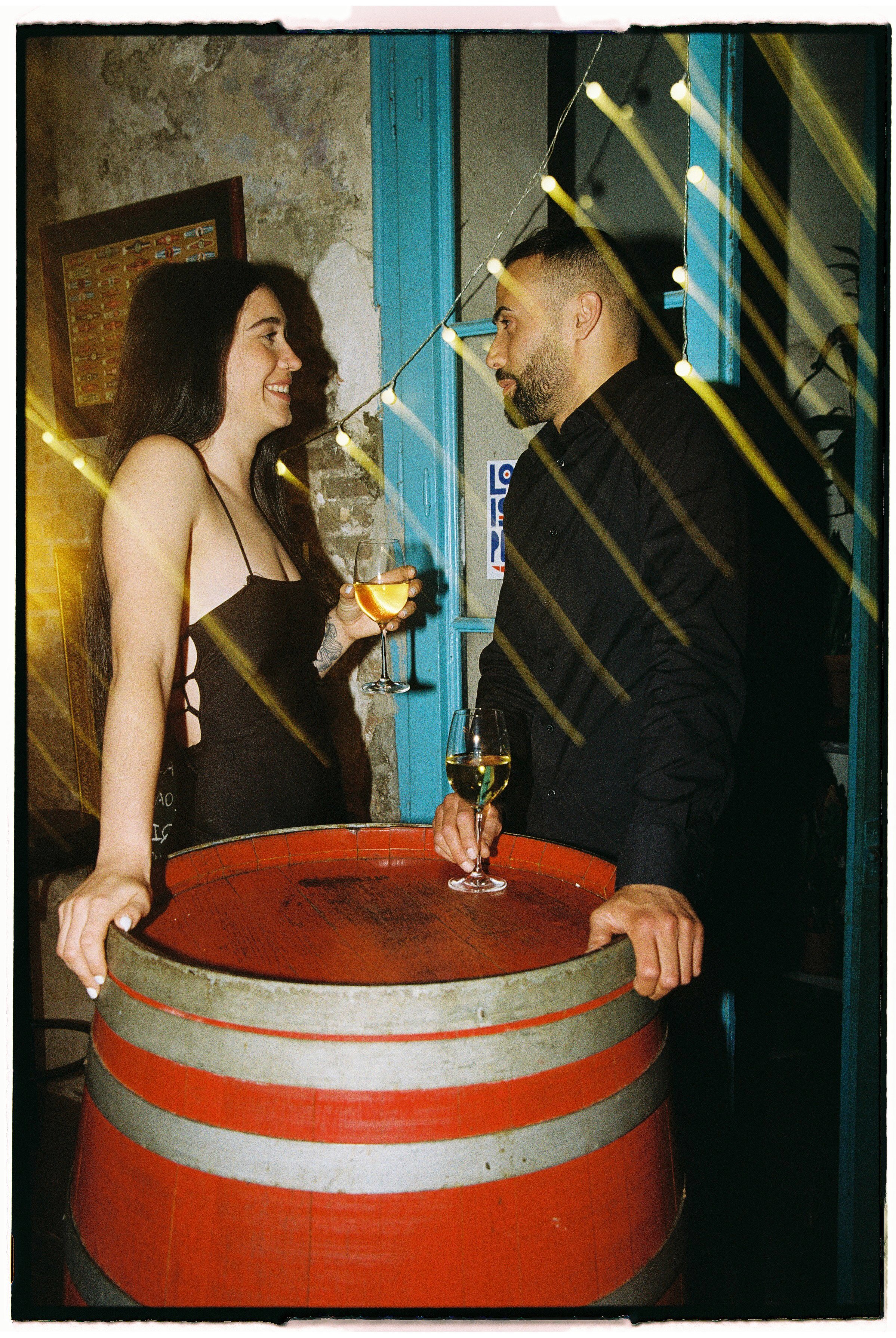 Una pareja en una celebración, sosteniendo copas de vino y charlando junto a una mesa que parece un barril de vino en un ambiente decorado con luces, paredes de piedra y un fondo de color turquesa.