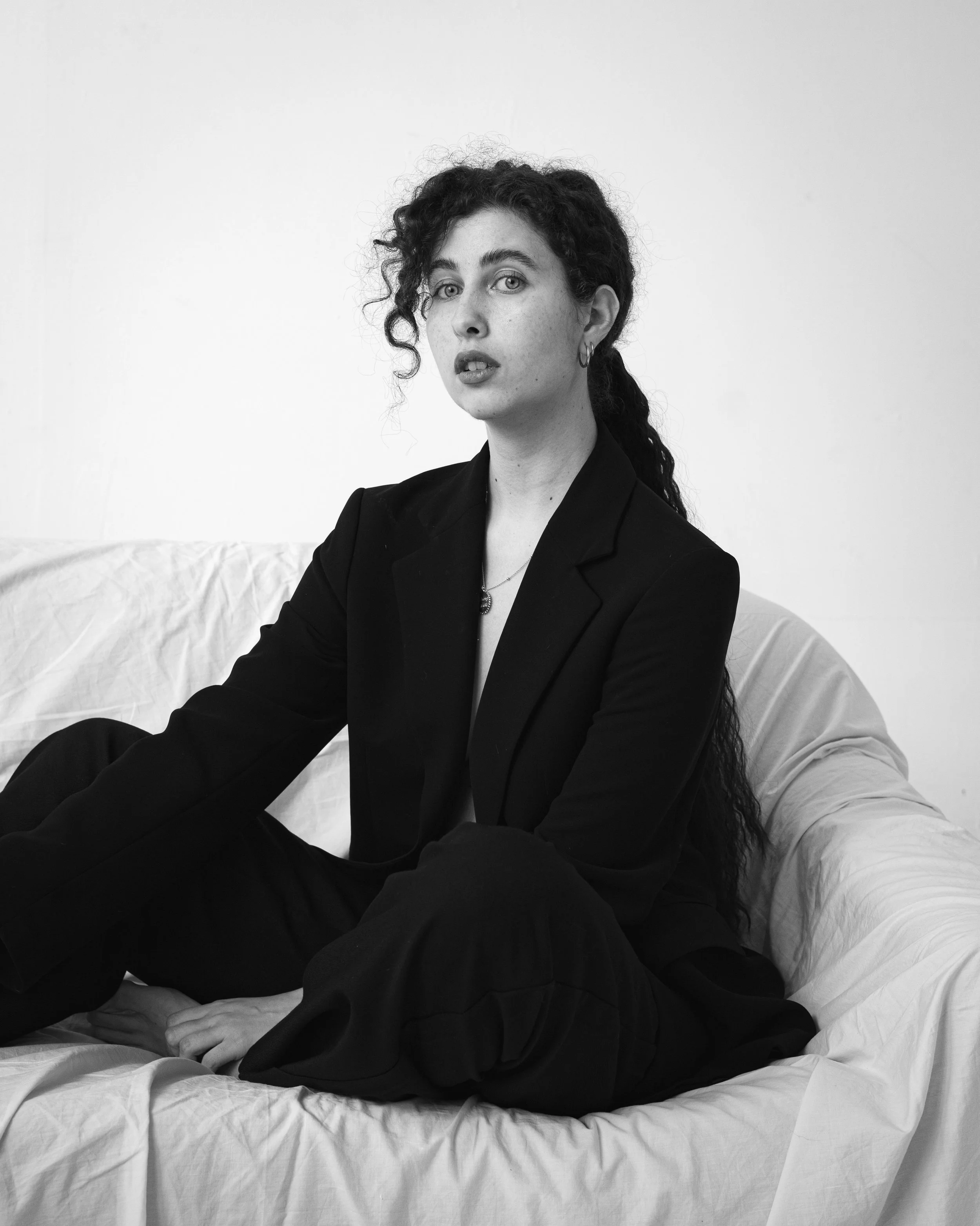 Retrato en blanco y negro de una mujer con cabello rizado, sin camisa, usando un blazer, sentada en un sofá con sábanas o mantas, en un fondo blanco.