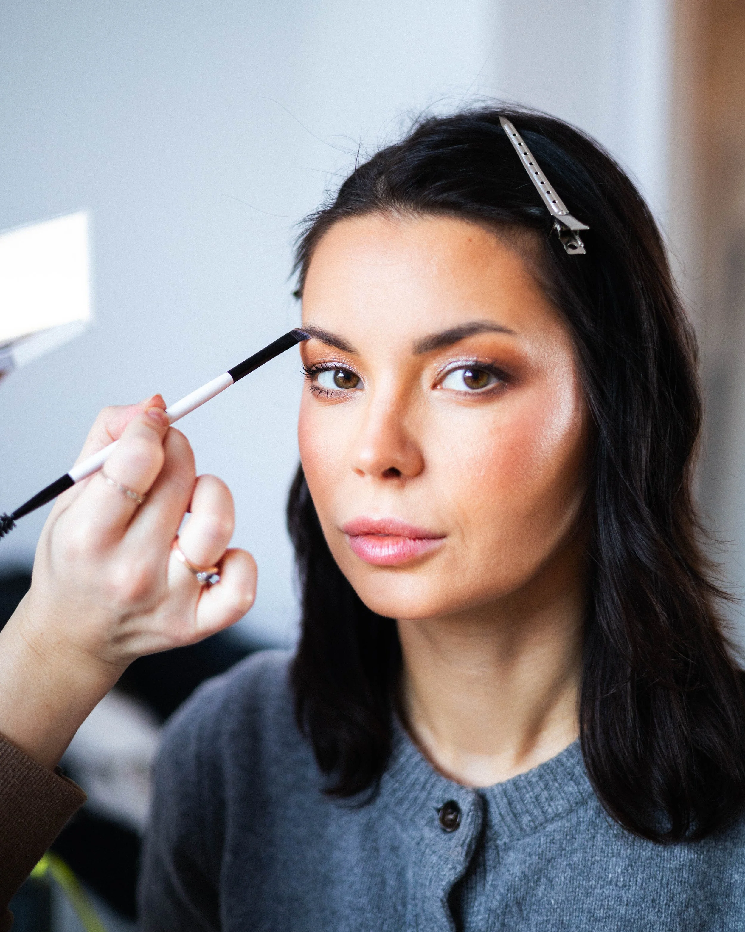 Una mujer con cabello oscuro se maquilla las cejas con un pincel en un ambiente bien iluminado.