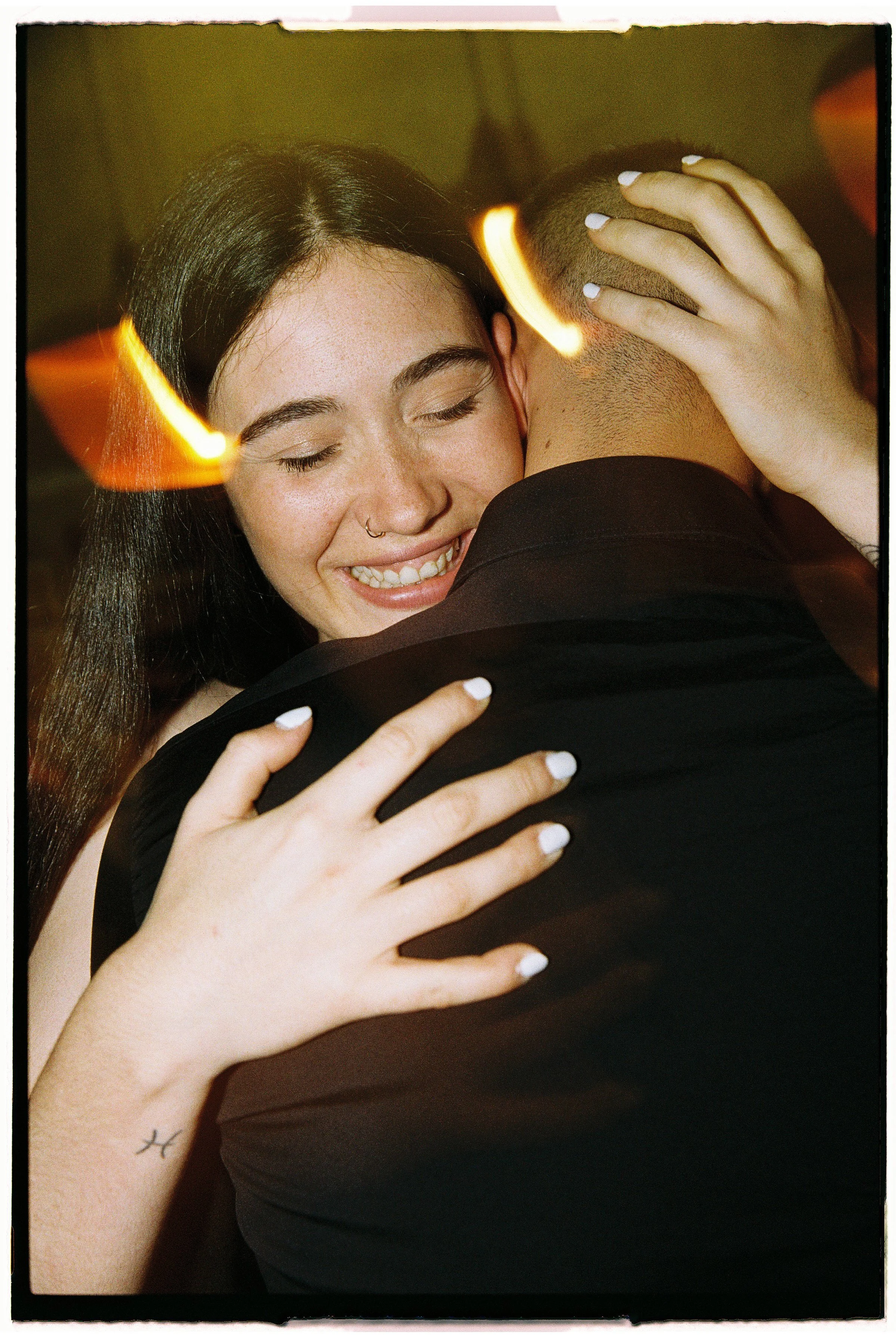 Una mujer y un hombre se abrazan con felicidad, la mujer tiene los ojos cerrados y una sonrisa, ella tiene el cabello largo y oscuro, y el hombre tiene la cabeza rapada y lleva puesta una chaqueta negra.