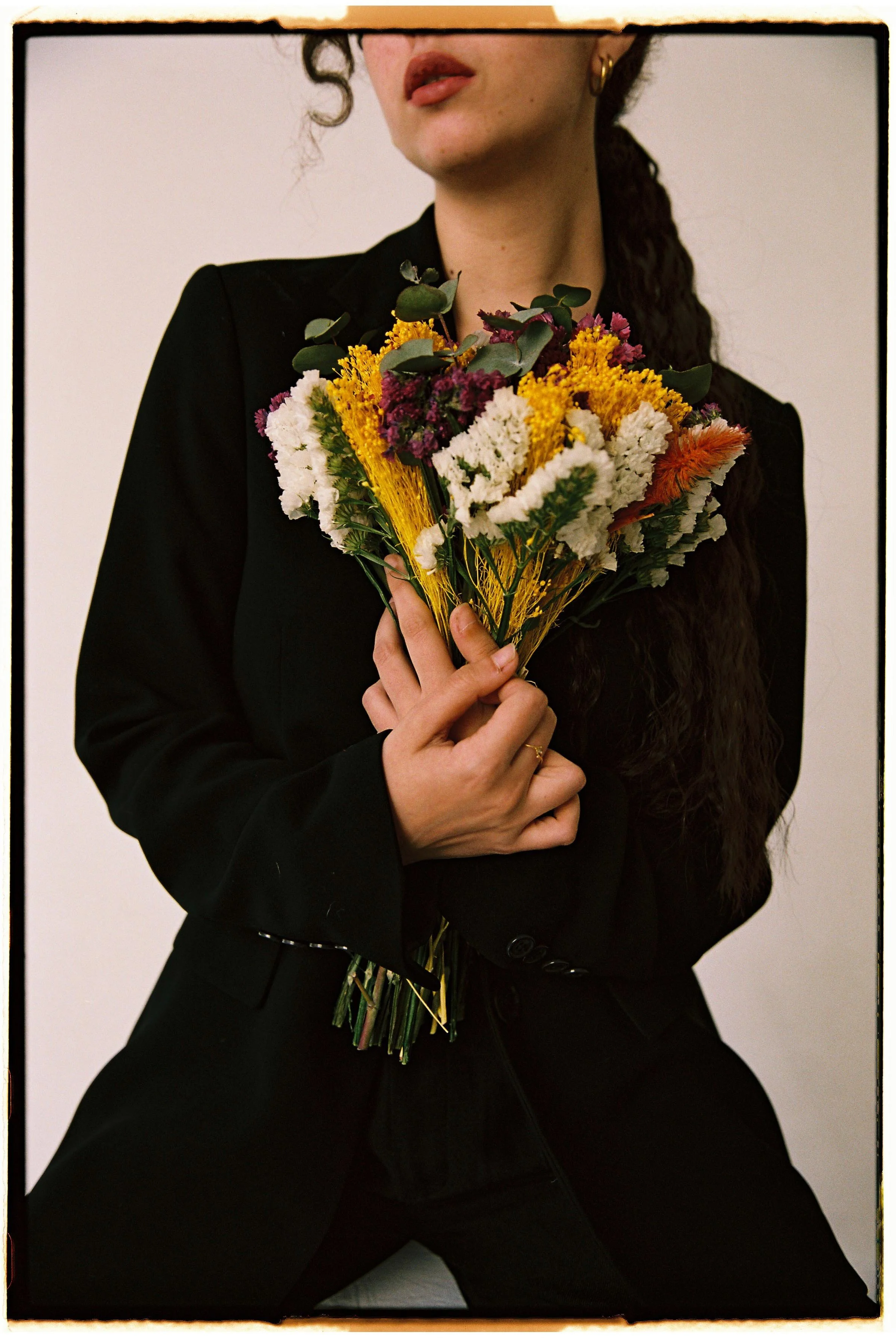 Persona con cabello largo y rizado, vestida de negro, sosteniendo un ramo de flores multicolores frente a un fondo neutro.