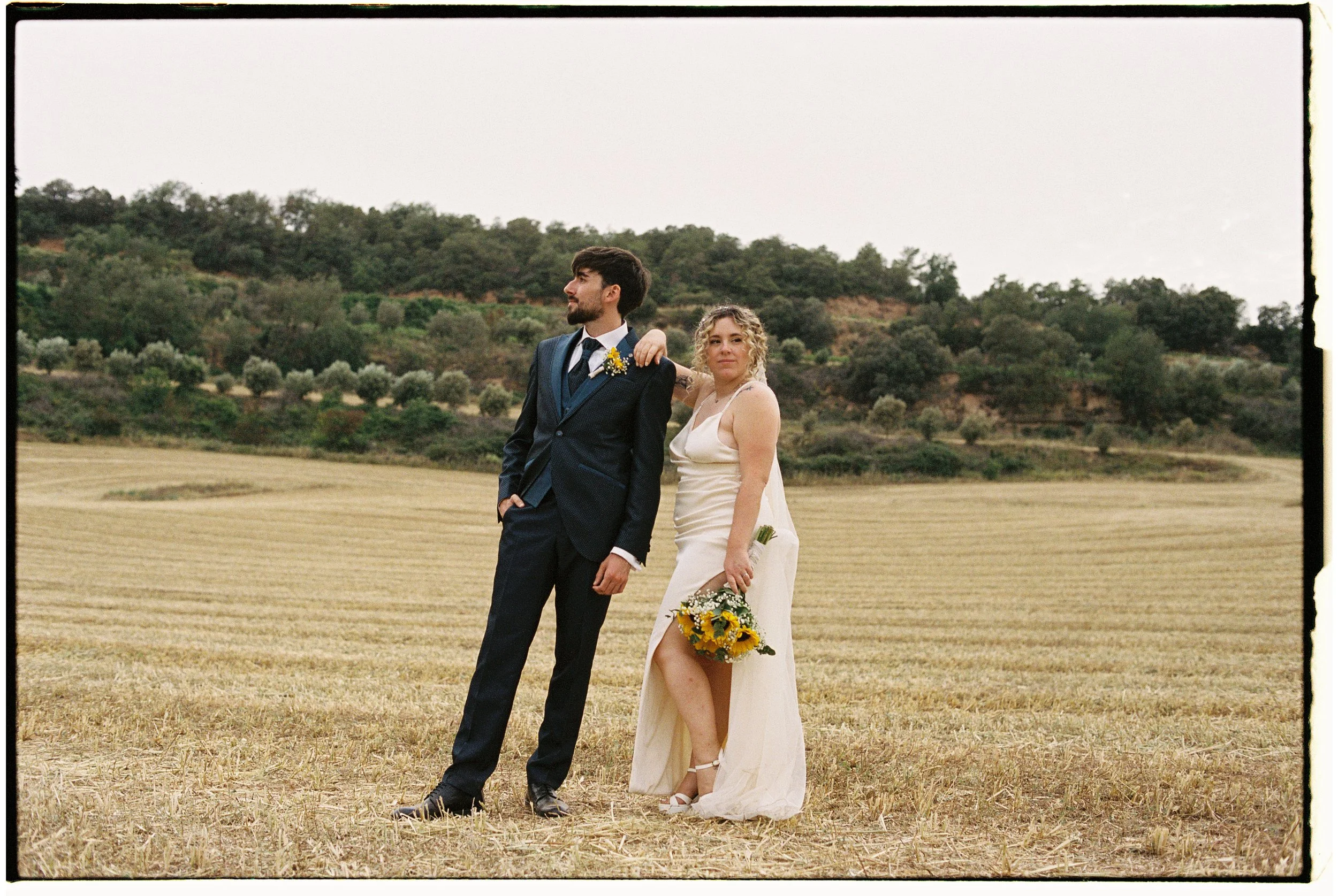 Una pareja de novios posando en un campo abierto, con un fondo de colinas y árboles, vestida con ropa de boda, la mujer sosteniendo un ramo de girasoles y flor de colores.