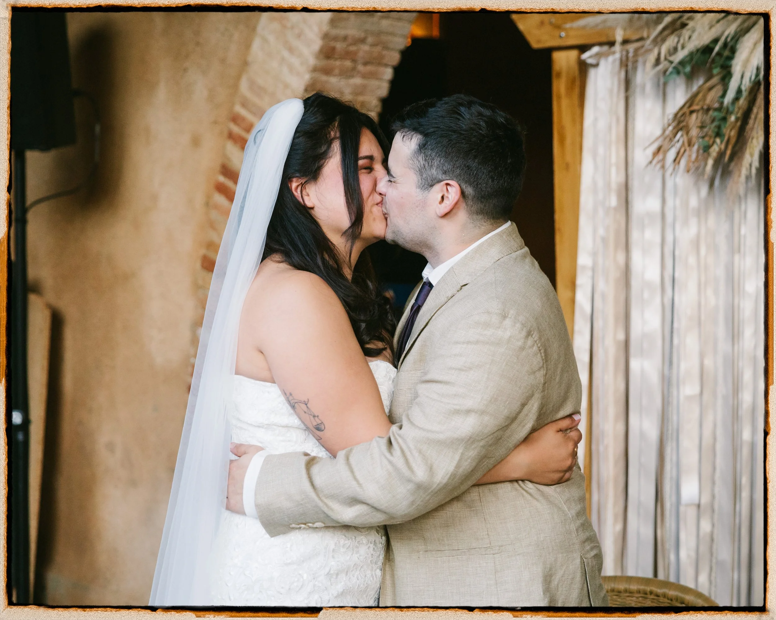 Pareja de recién casados besándose con los ojos cerrados, en un ambiente rústico con paredes de ladrillo y cortinas transparentes.