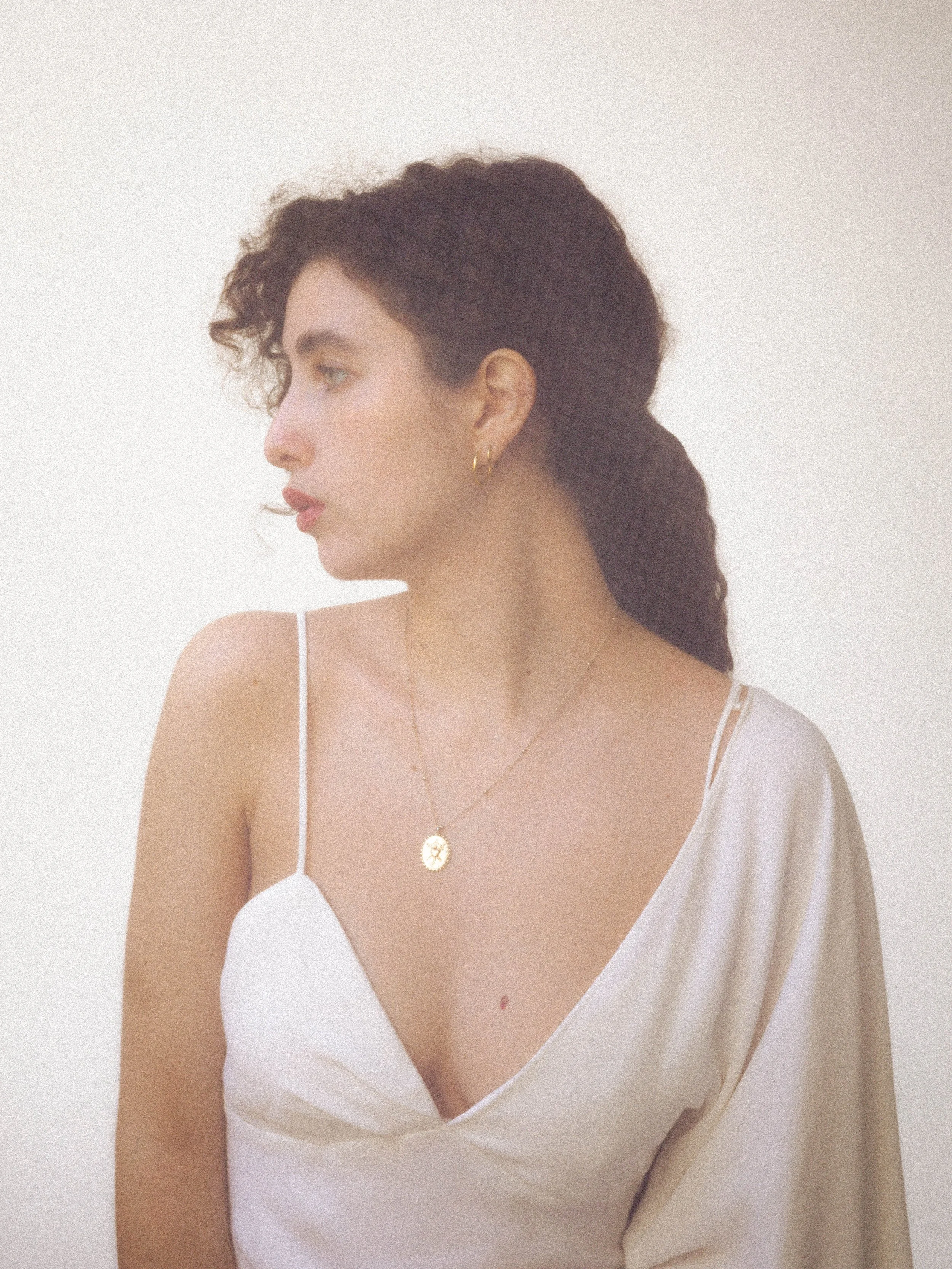 Retrato de una mujer de perfil con cabello rizado oscuro, usando collar dorado y una blusa blanca con tirantes, contra un fondo blando.