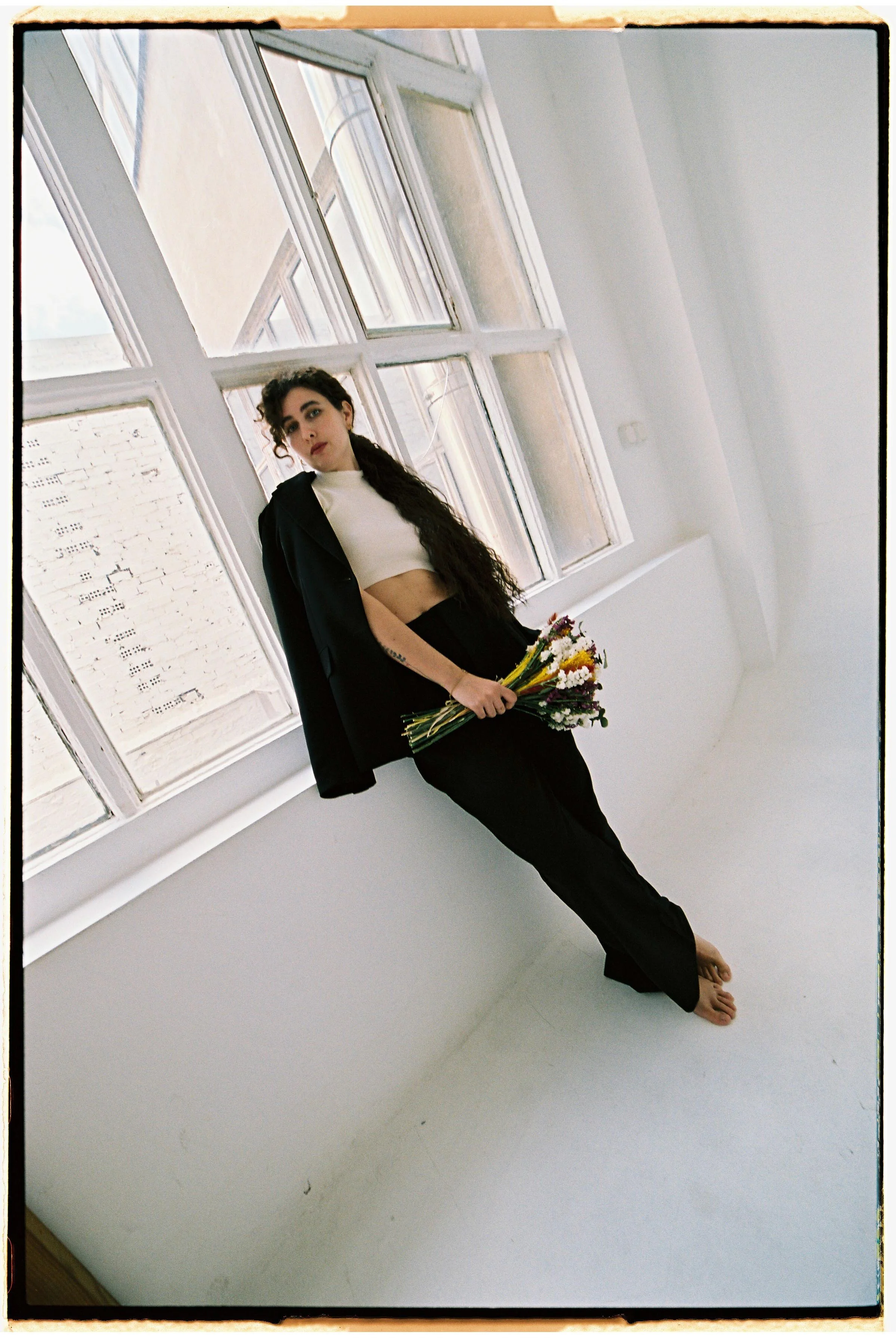 Una mujer con cabello largo y oscuro, vestidos negro y blanco, sosteniendo un ramo de flores, en una habitación con grandes ventanas y paredes blancas.