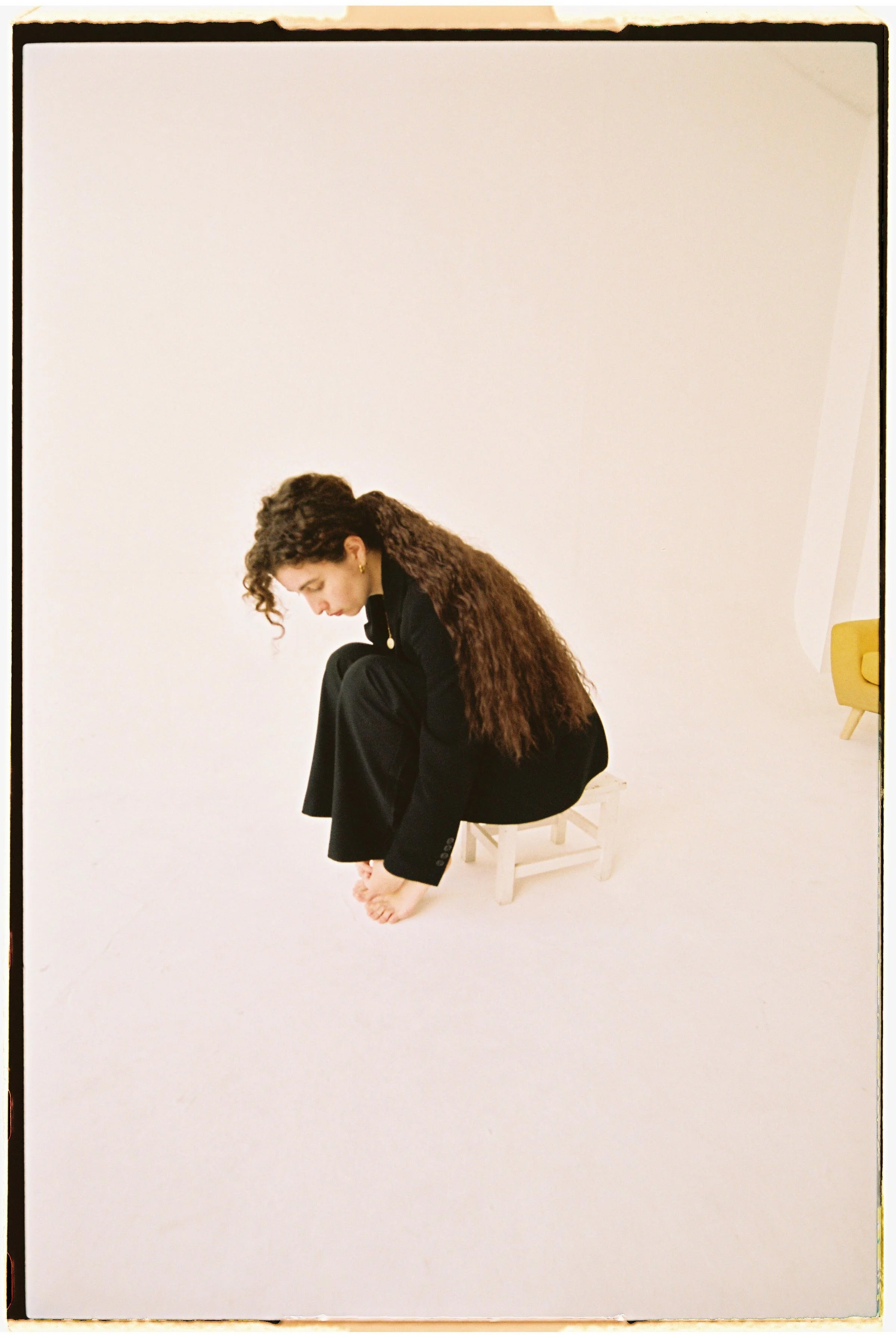 Mujer sentada en una silla diminuta en un fondo blanco, con cabello rizado y cabello largo, vestida con traje oscuro, en posición de doblar sus piernas y tocarlas con las manos, bebiendo agua.