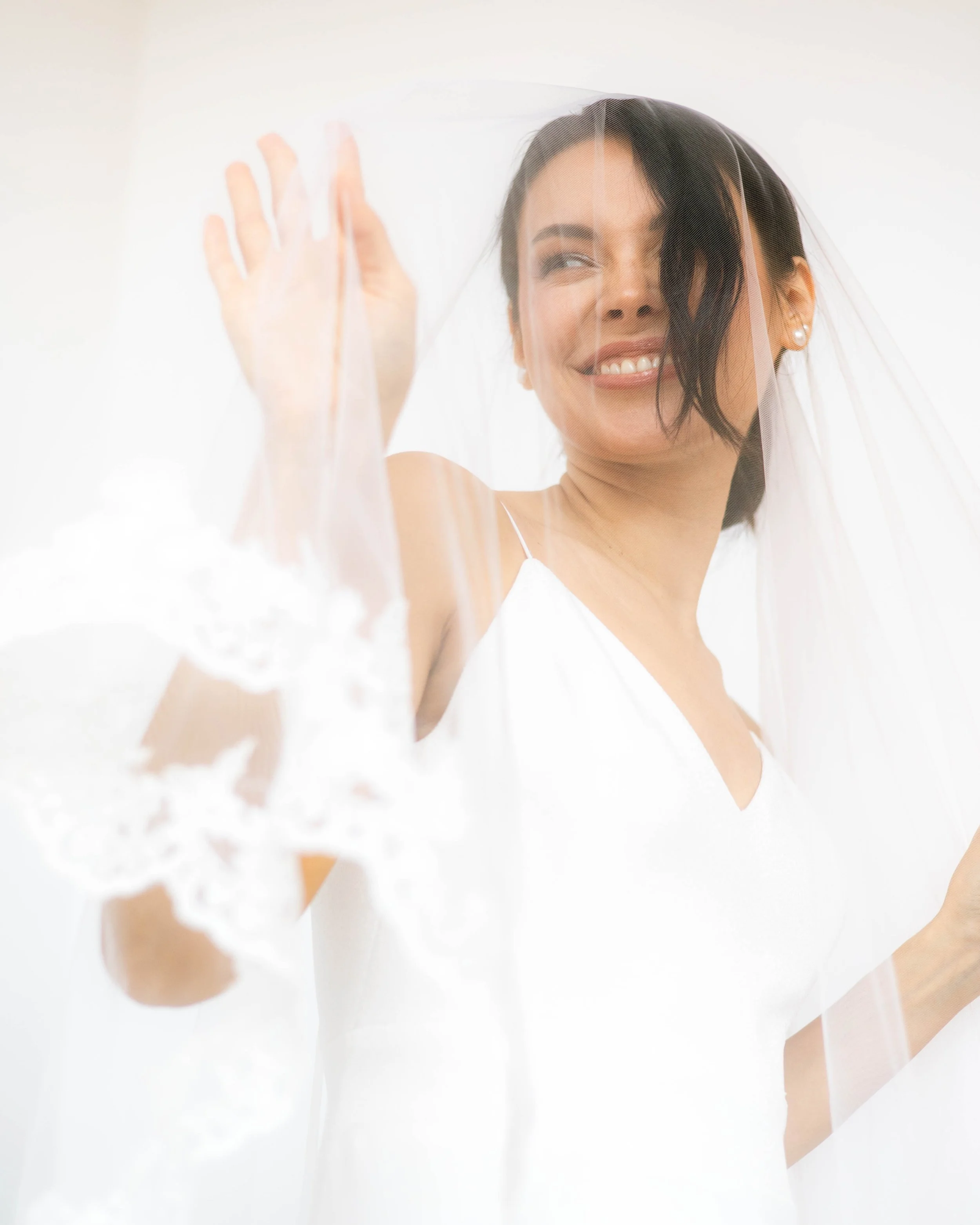 Mujer con vestido de novia y velo, sonriendo y tocando una cortina de encaje.