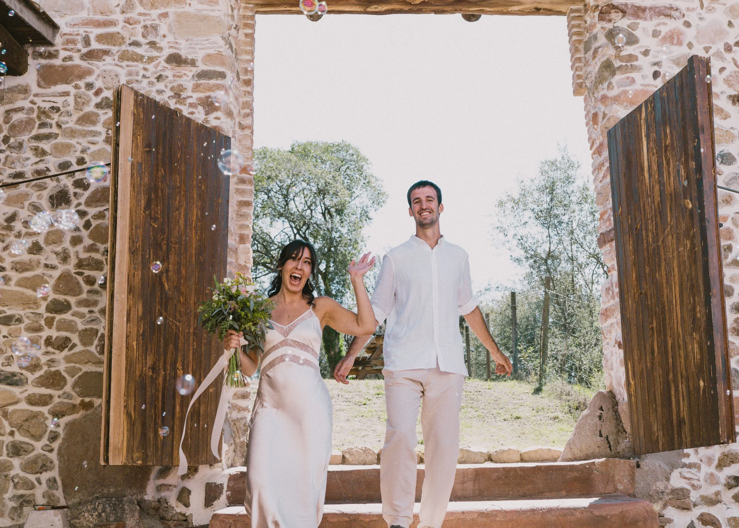 Pareja de recién casados saliendo de una iglesia rústica con puertas de madera, celebrando y sonriendo, rodeados de árboles y flores.