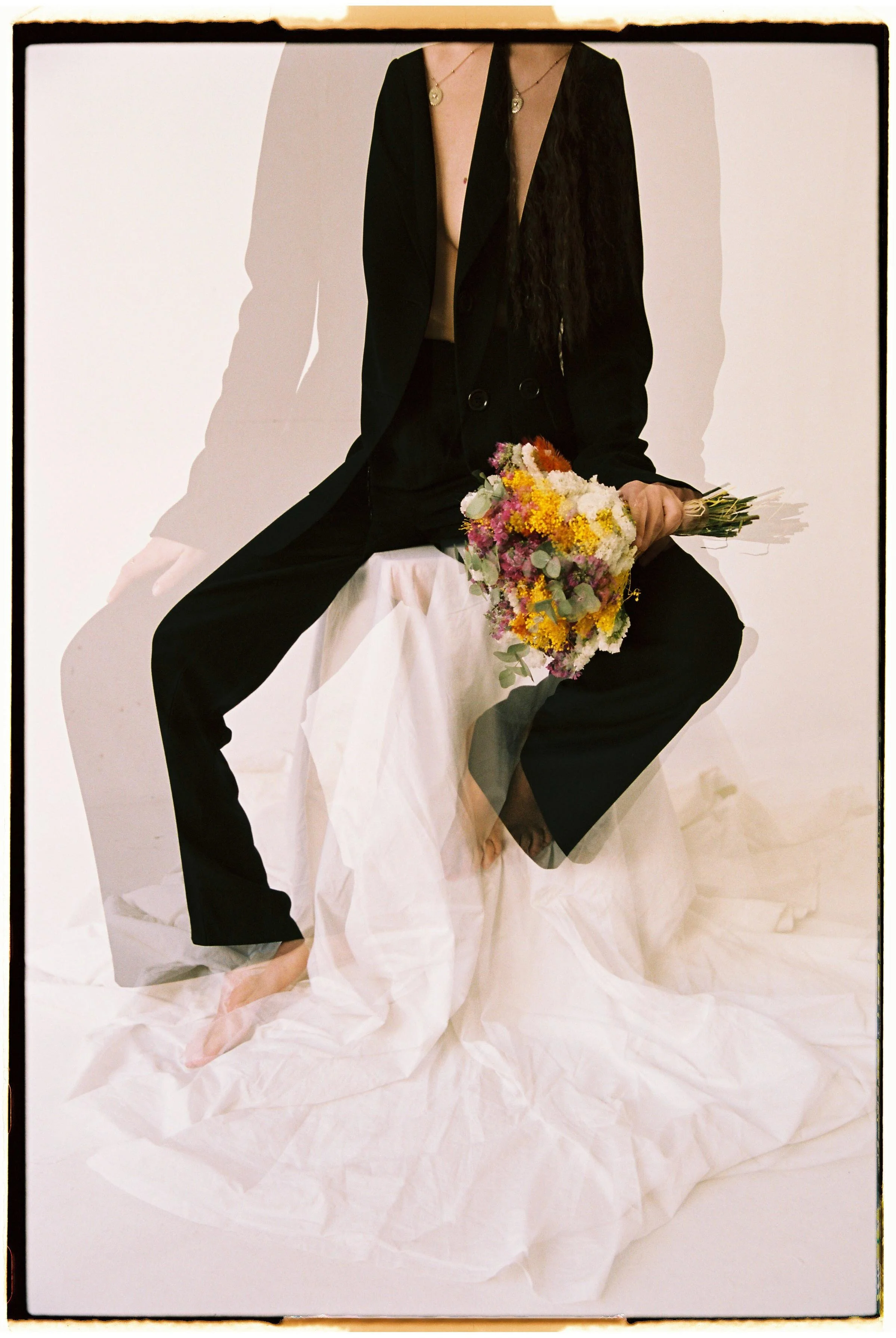 Persona sentada con traje negro y sin blusa, sosteniendo un ramo de flores coloridas, sobre un fondo blanco y con telas blancas en el suelo.