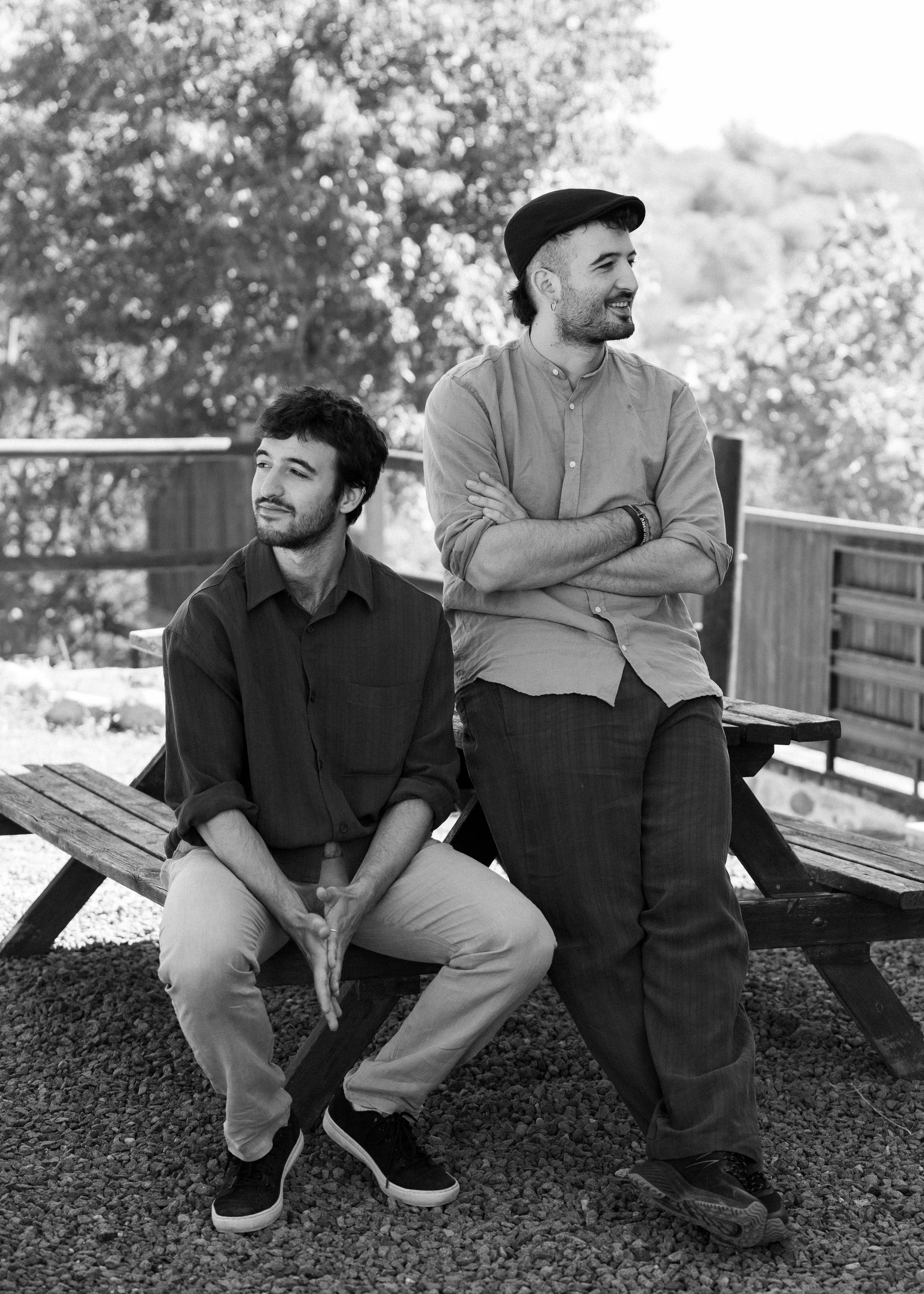 Dos hombres en un parque, uno sentado sobre un banco de madera con las manos juntas y otro de pie con los brazos cruzados, ambos sonriendo y conversando al aire libre.