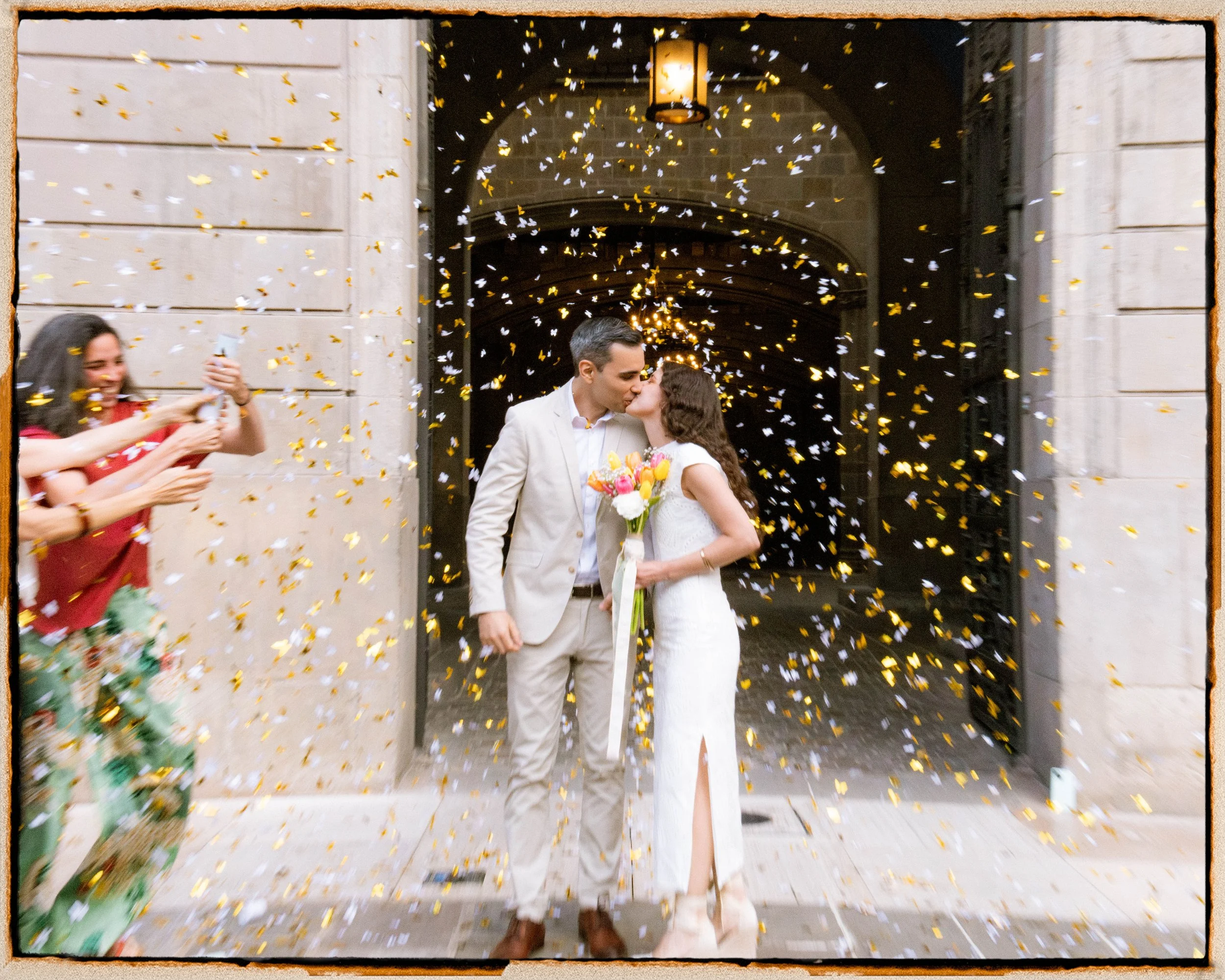 Una pareja de recién casados besándose en la entrada de un edificio, rodeados de confeti dorado y blanco, mientras otros celebrantes toman fotos.