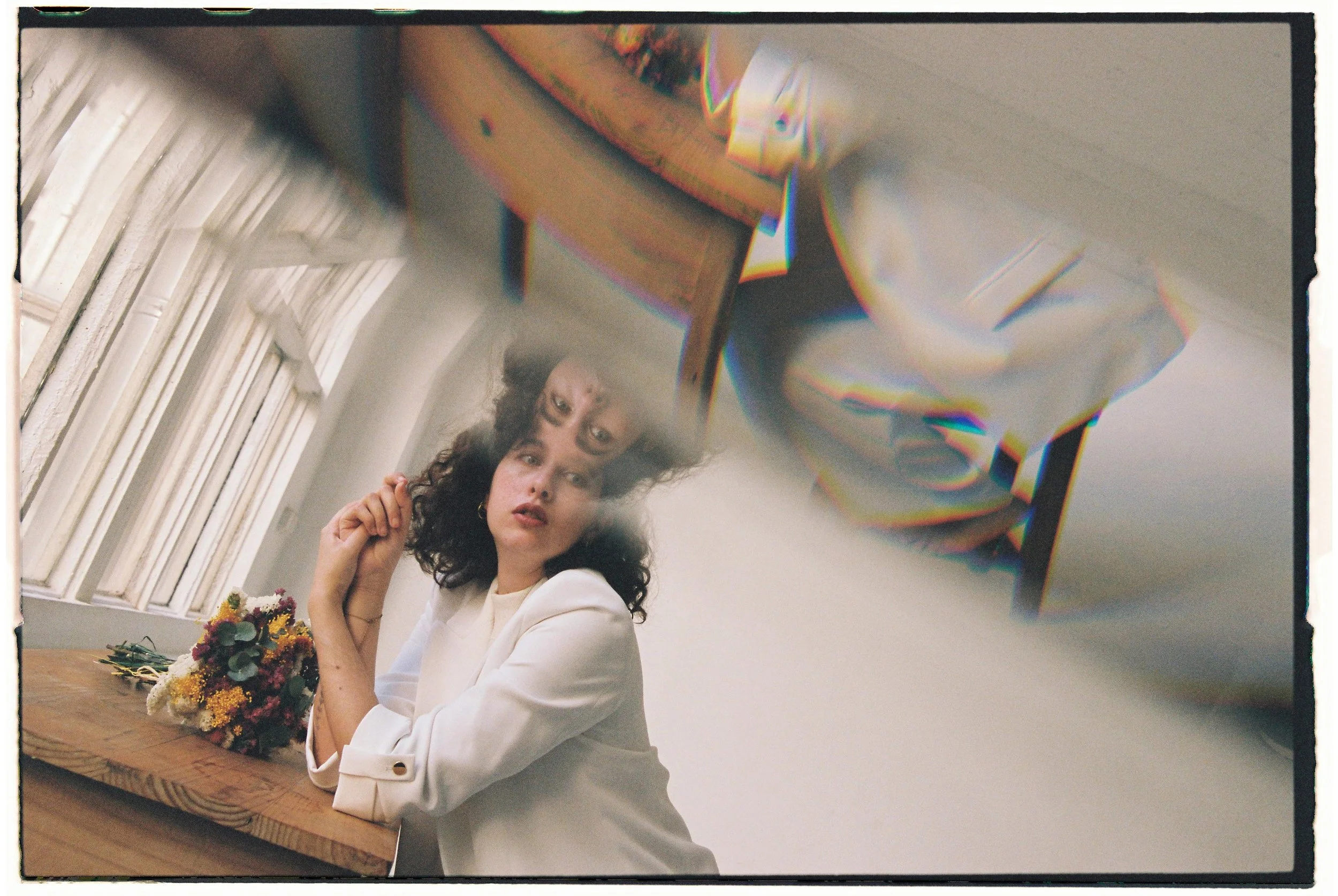 Una mujer con cabello rizado y vestido blanco, sentada en una mesa de madera con un ramo de flores, mira hacia la cámara en una habitación con ventanas y un reflejo en un espejo.