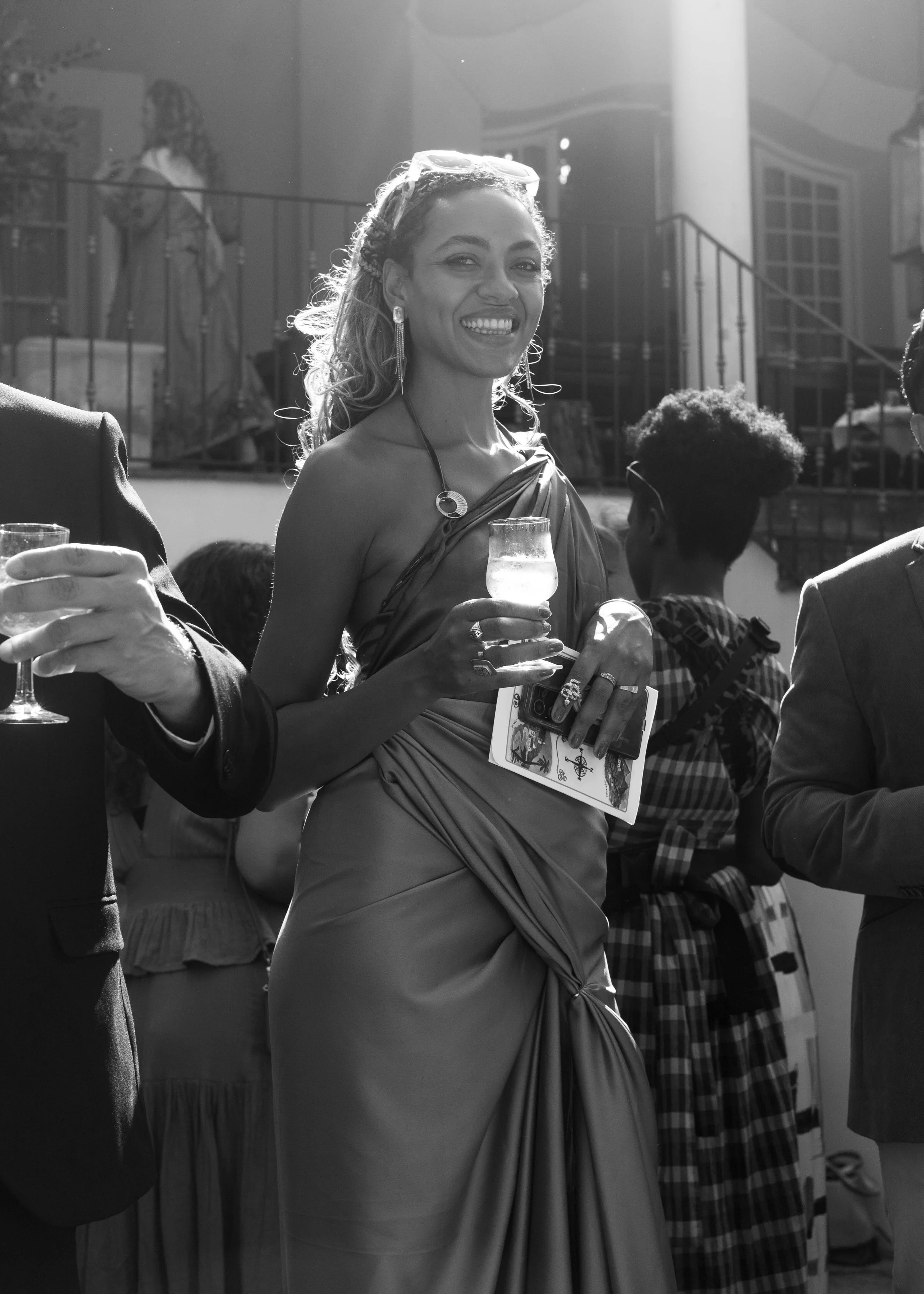 Mujer sonriendo, vestida con elegante vestido y accesorios, sosteniendo una copa en un evento social, en un ambiente con otras personas y escaleras en el fondo.