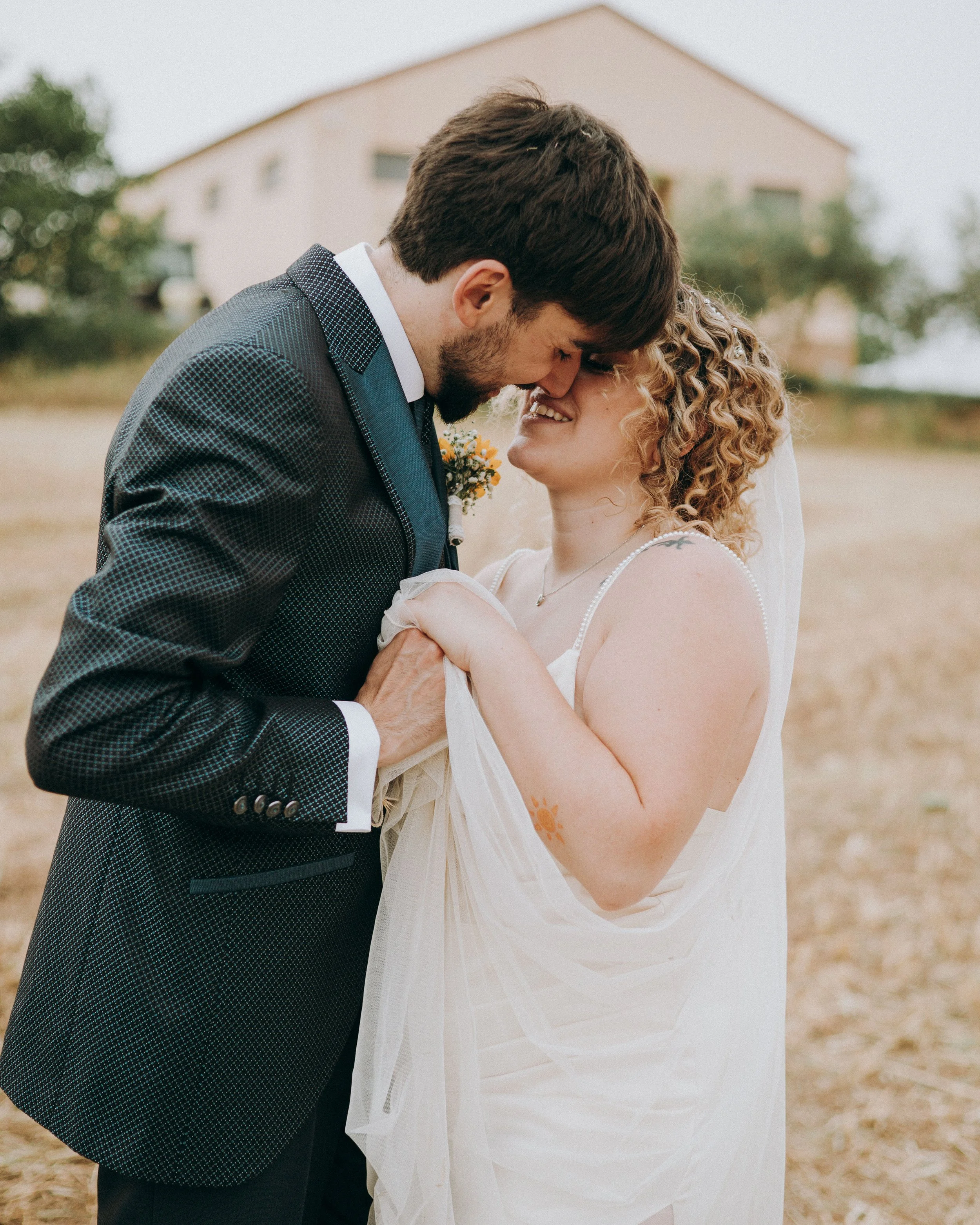 Pareja de recién casados en una pose romántica en un campo, con la frente tocándose y sonriendo, ella con vestimenta blanca y el con traje oscuro.