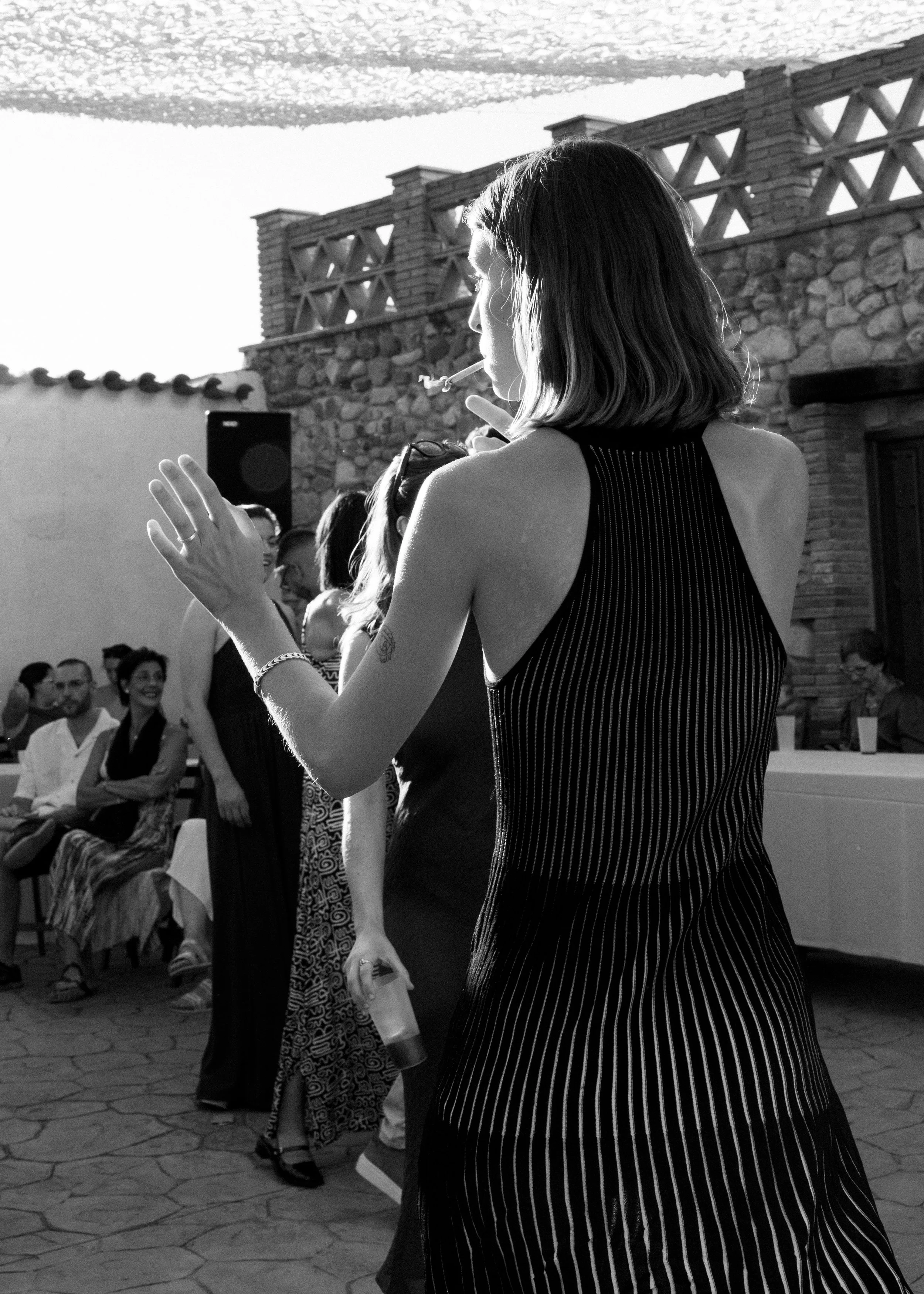 Mujer joven bailando en una fiesta al aire libre, con fondo de personas sentadas y una pared de piedras.