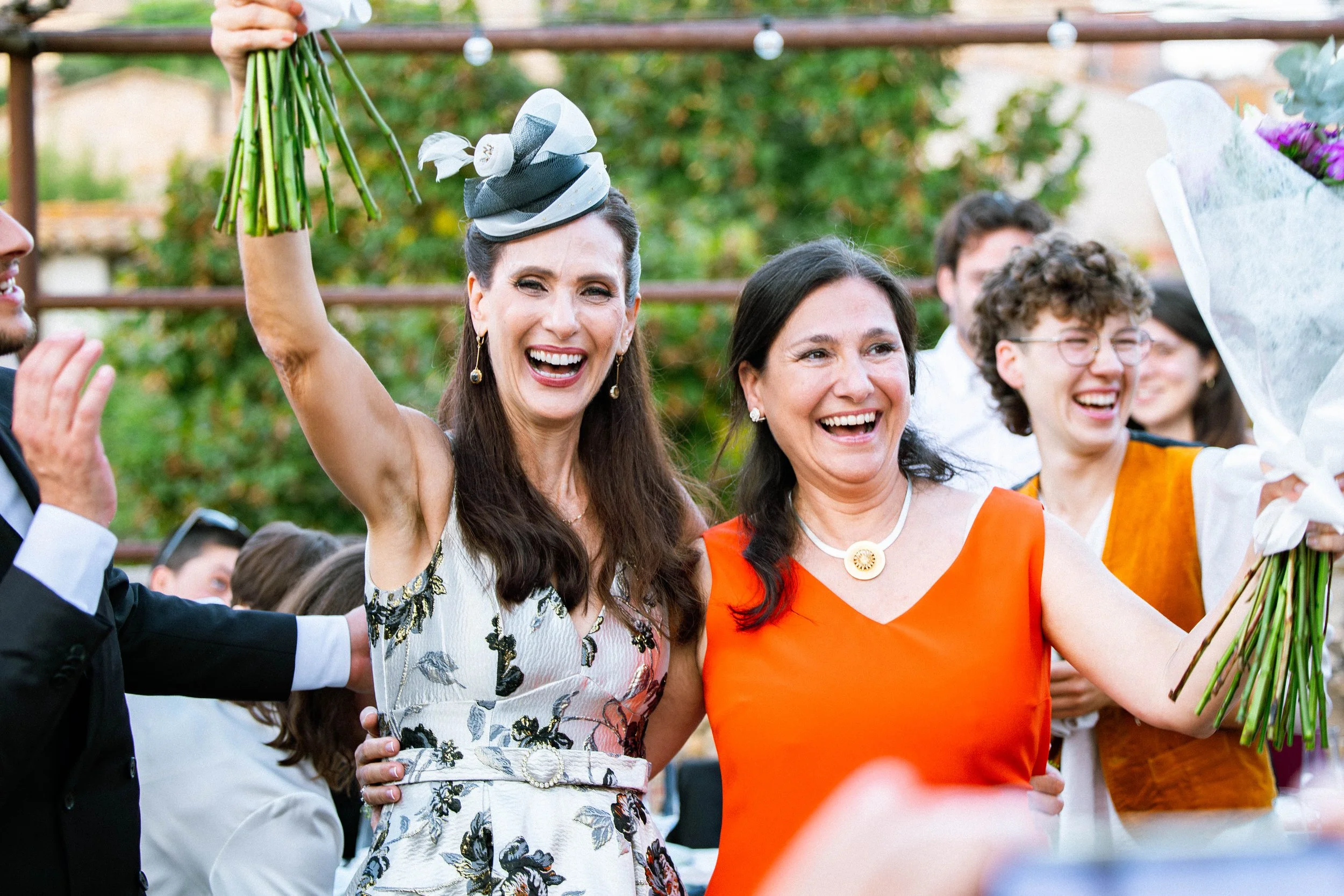 Grupo de personas en una celebración, con una mujer en el centro sosteniendo un ramo de flores y sonriendo, rodeada de otras personas que también están felices y usando ropa de colores brillantes.