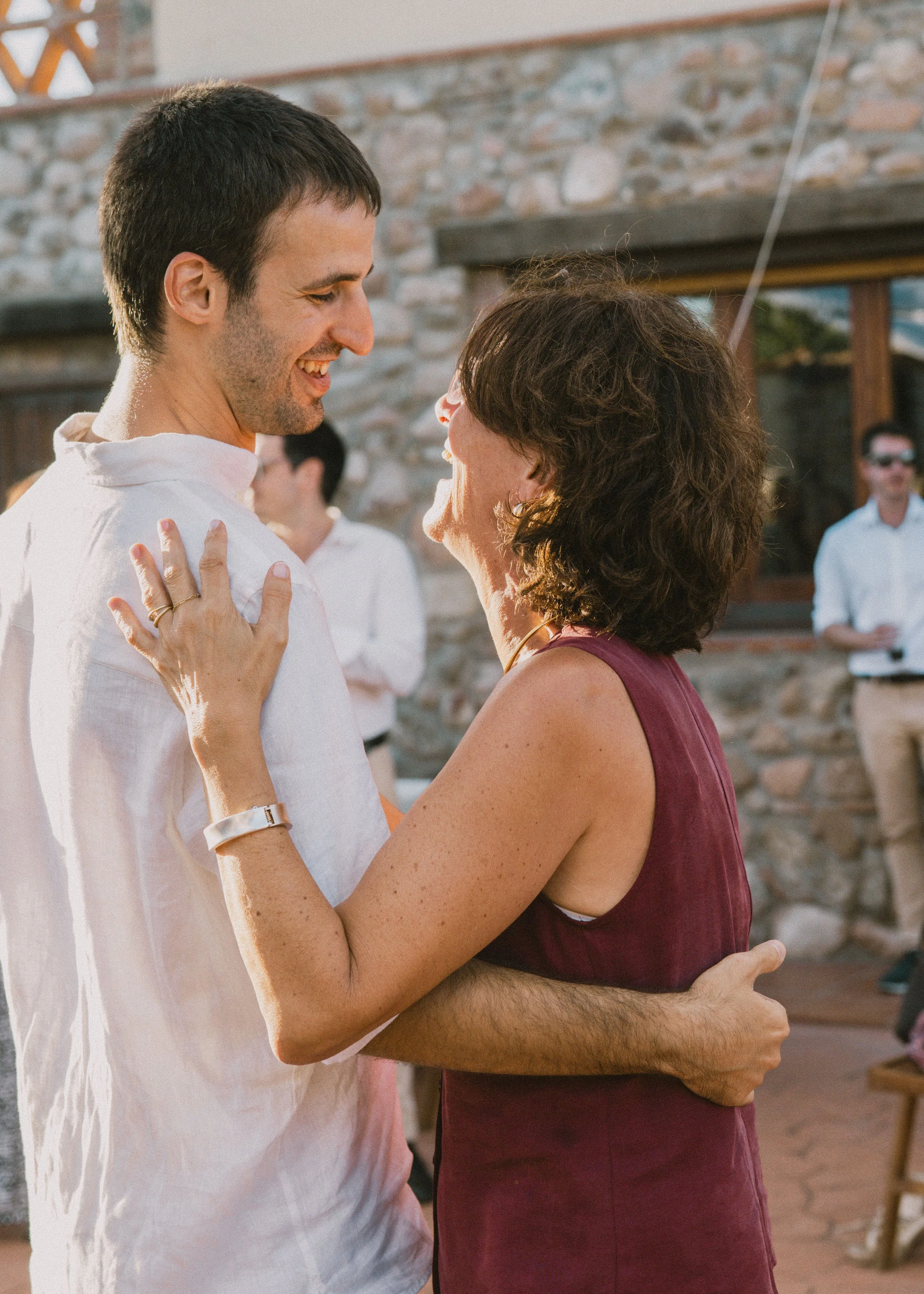 Un hombre y una mujer se están abrazando y sonriendo en una reunión al aire libre, con un fondo de pared de piedra y otros invitados en el fondo.