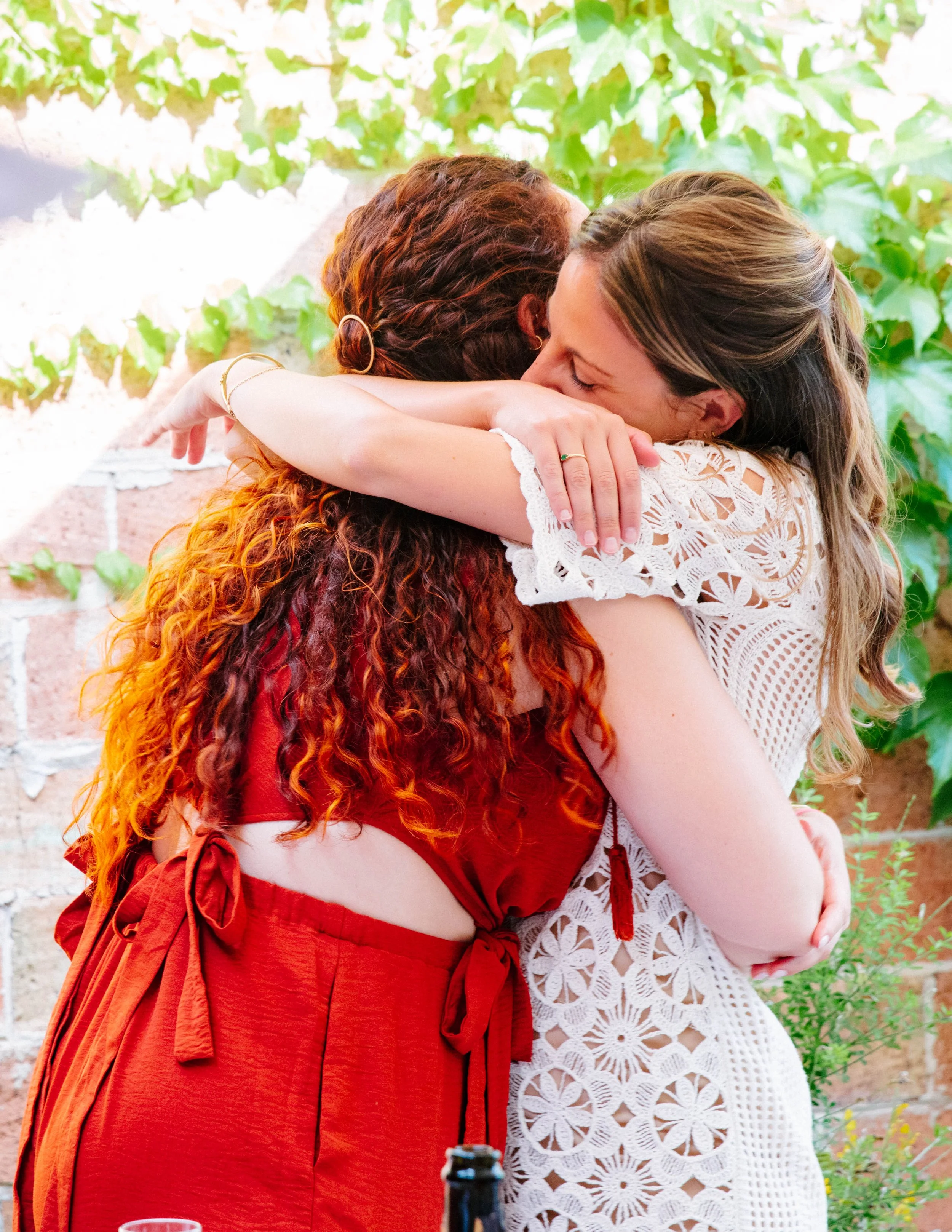 Dos mujeres se abrazan afectuosamente en un entorno natural con plantas verdes en el fondo.