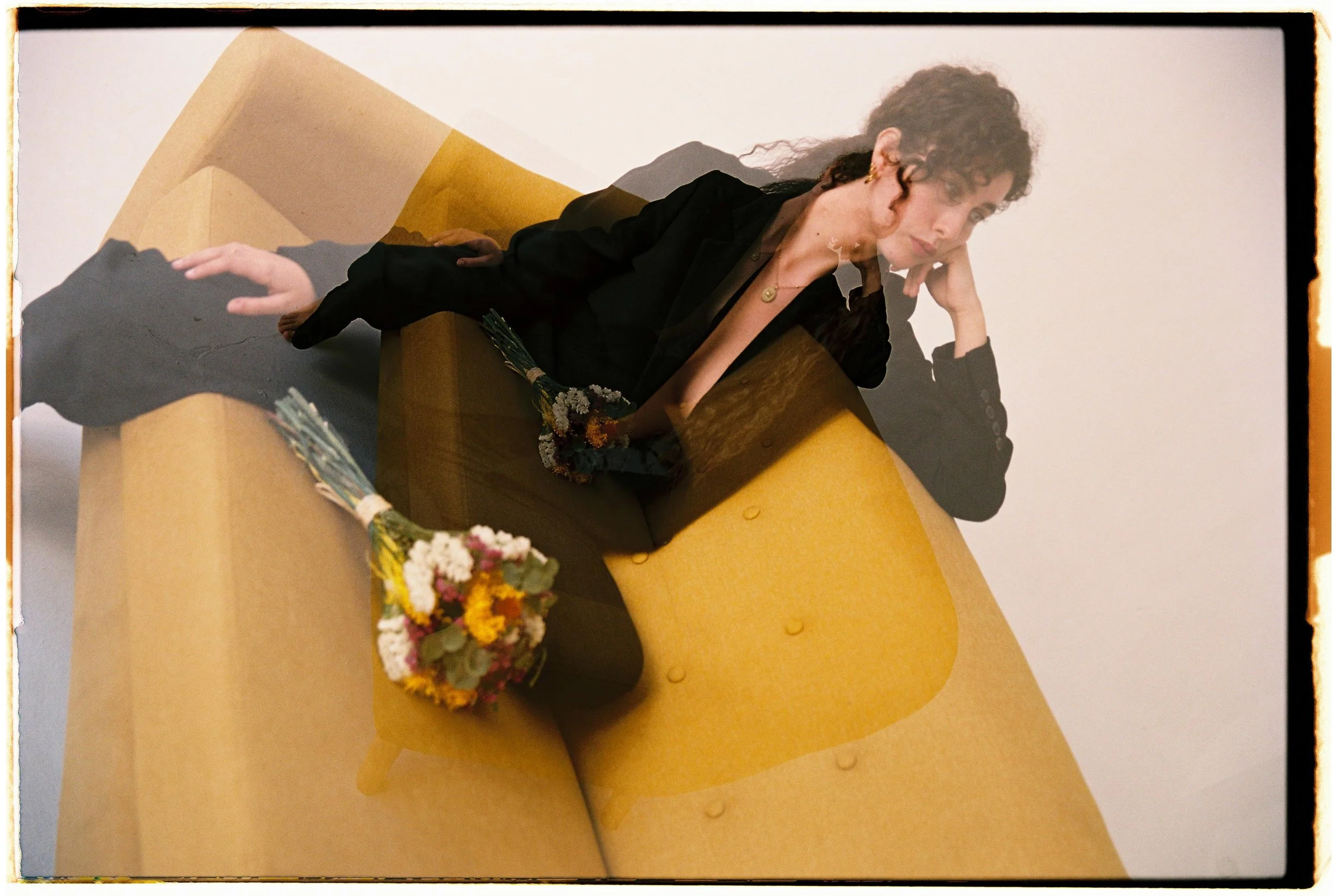 Mujer de cabello rizado y piel clara, vestida con saco negro, recostada en sillón amarillo, con un ramo de flores sobre las piernas.