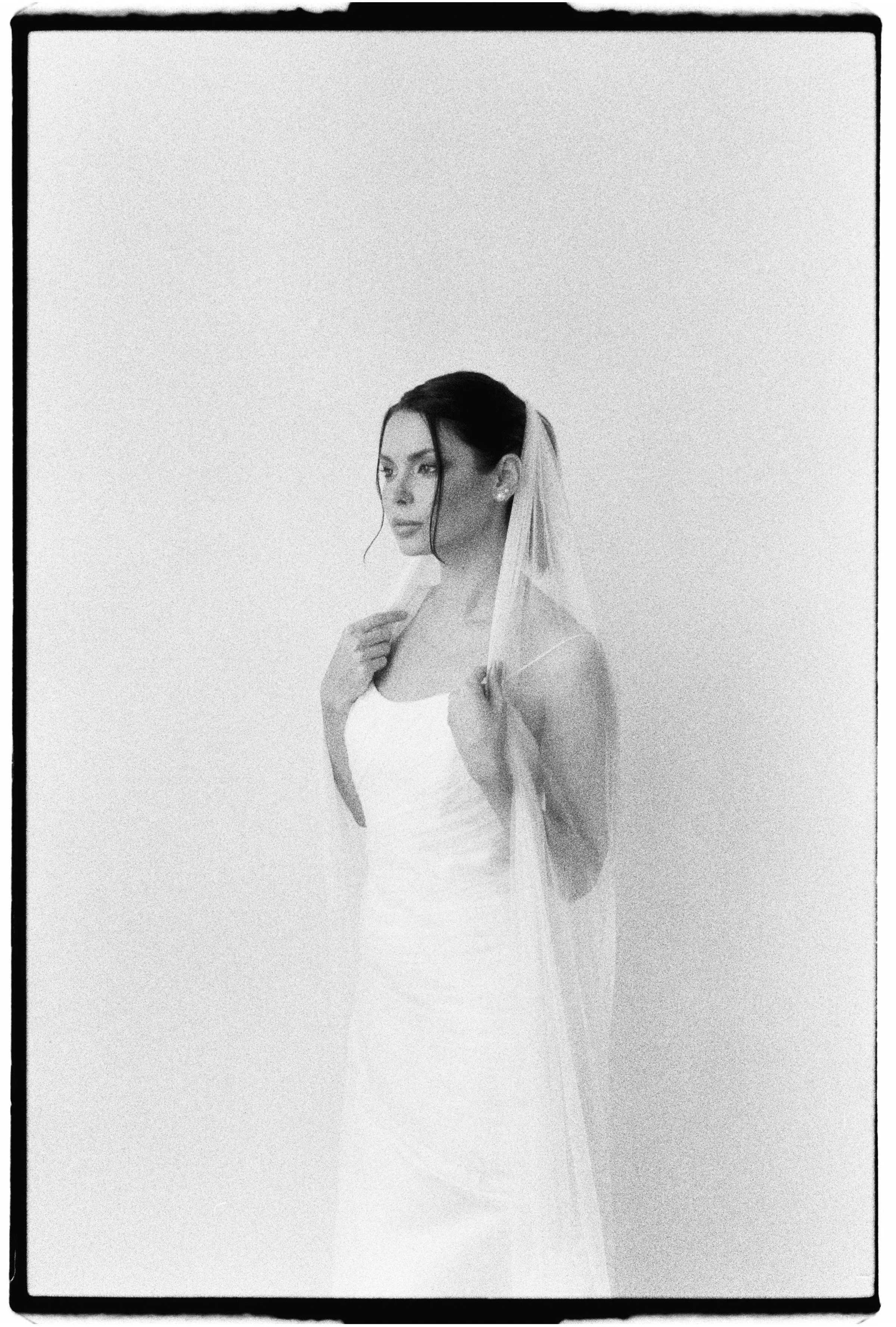 Mujer con vestido de novia y velo, de perfil, posando contra una pared de fondo claro.