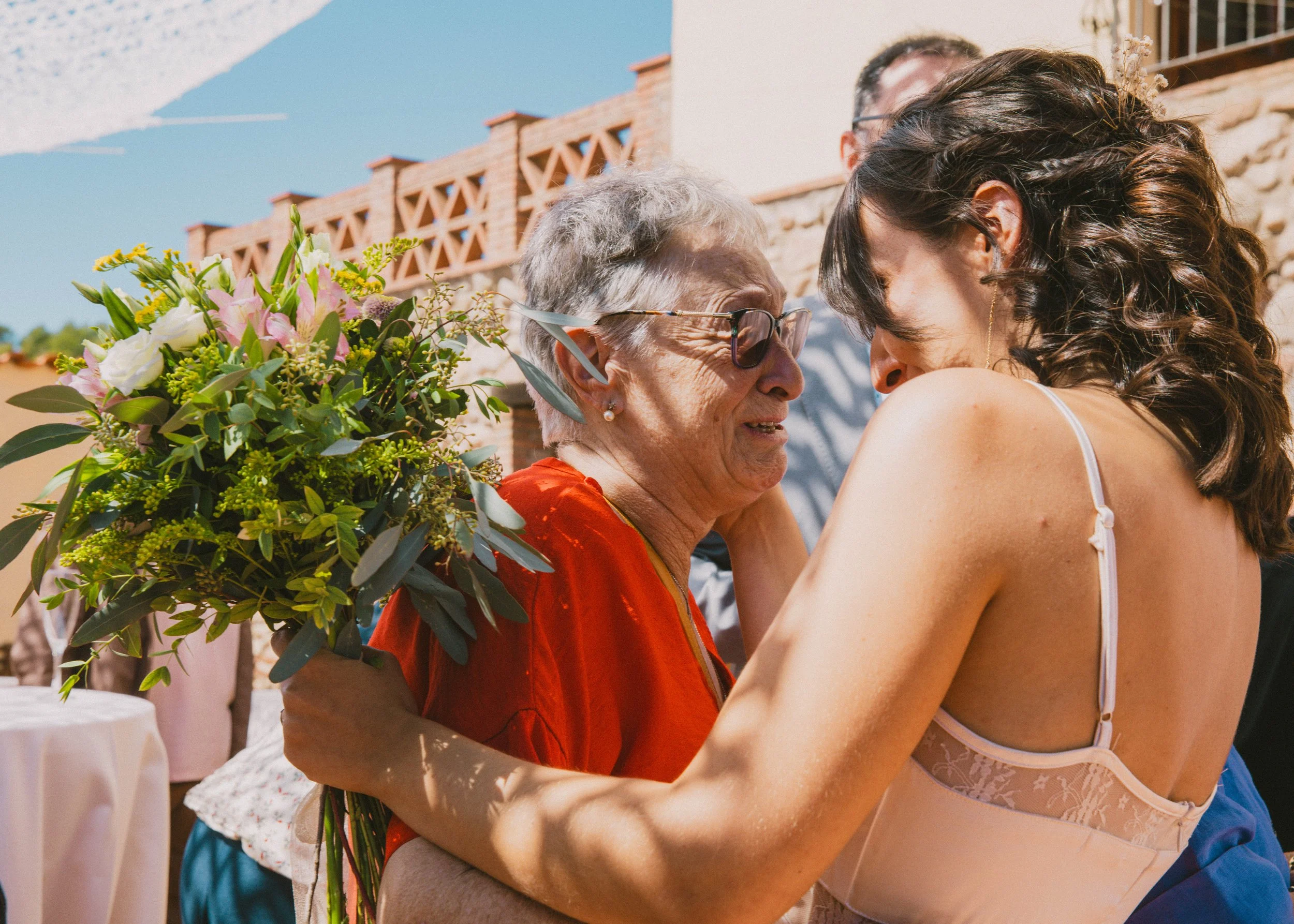 Una joven y una anciana se abrazan y sonríen en una reunión al aire libre, la joven le entrega un ramo de flores a la anciana, en un día soleado.