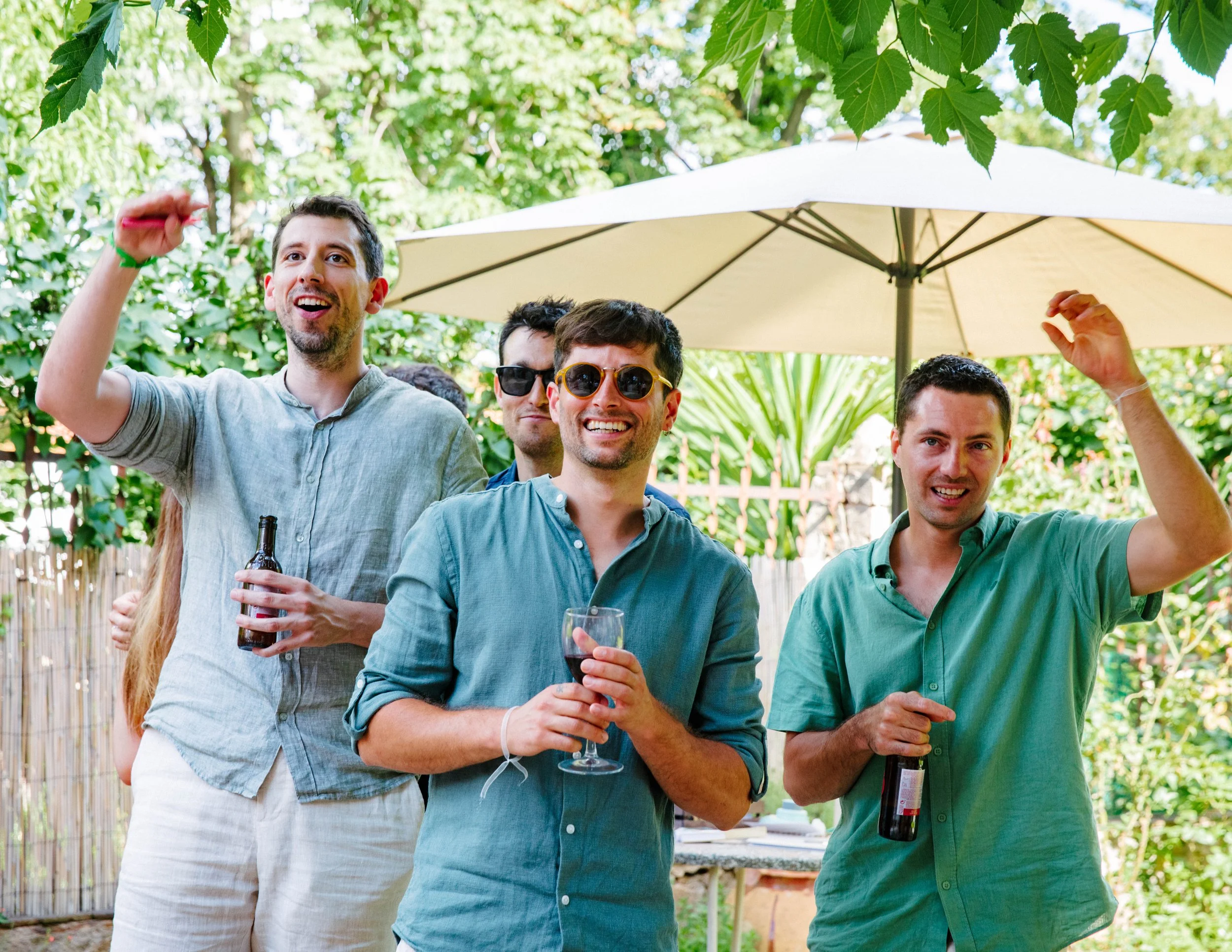Grupo de amigos en una reunión al aire libre, algunos con gafas y sosteniendo botellas de vino y una copa de vino, en un entorno con árboles y sombrillas.