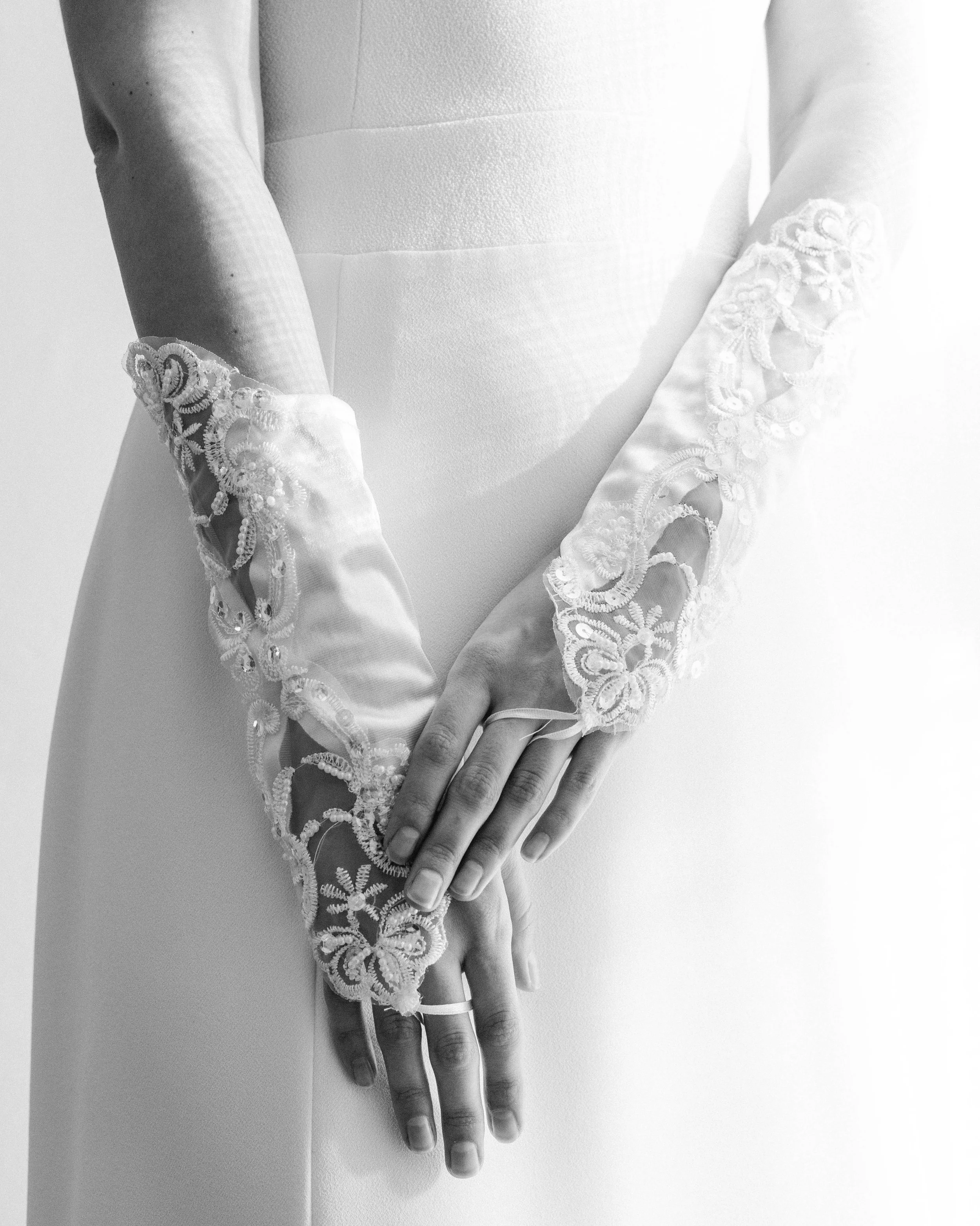 Manos de una novia con guantes de encaje mirando hacia abajo, la mano sostiene un anillo, vestido de boda blanco.