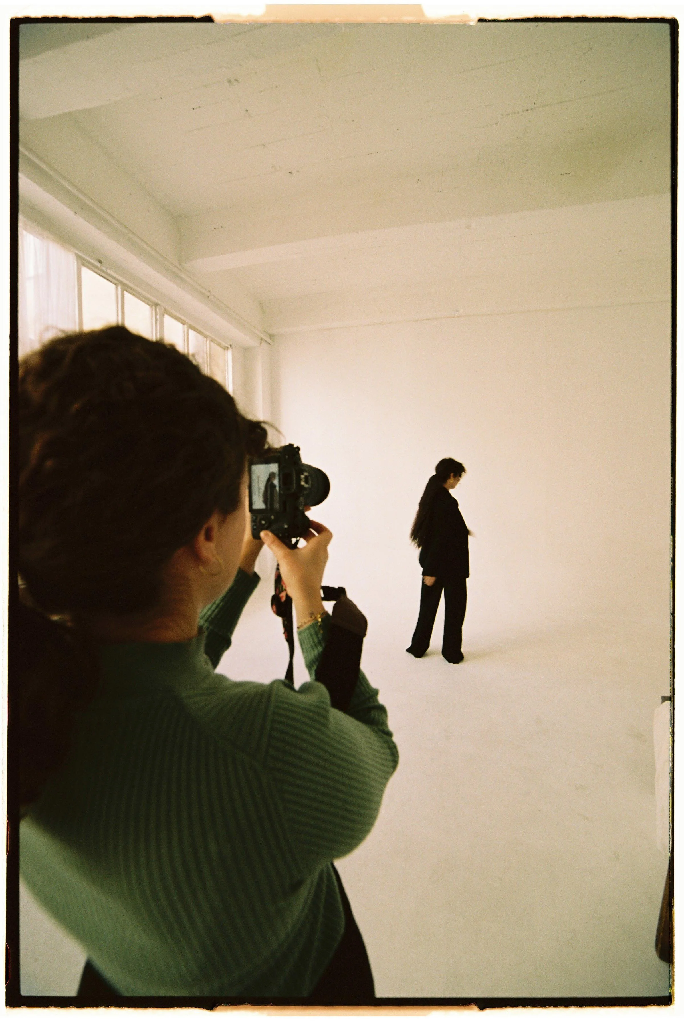 Una mujer toma una foto de otra mujer de espaldas en un espacio grande y vacío, con paredes blancas y ventanas largas a la izquierda.