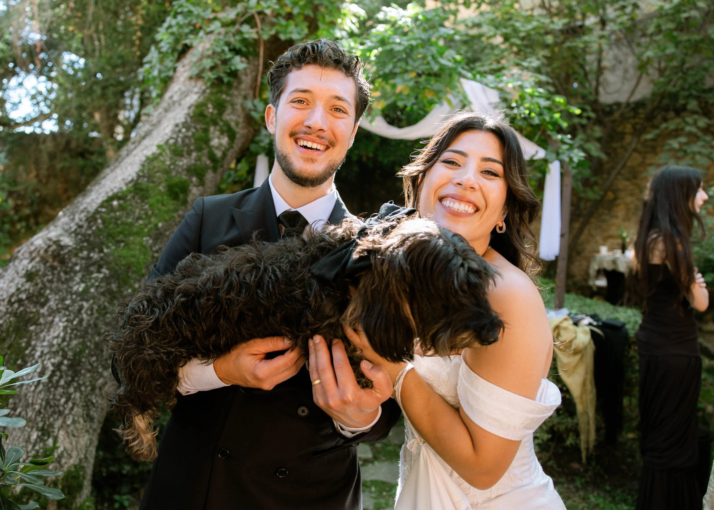 Pareja en celebración de boda feliz, abrazándose con perros pequeños en brazos, rodeados de árboles y decoración en un espacio al aire libre.