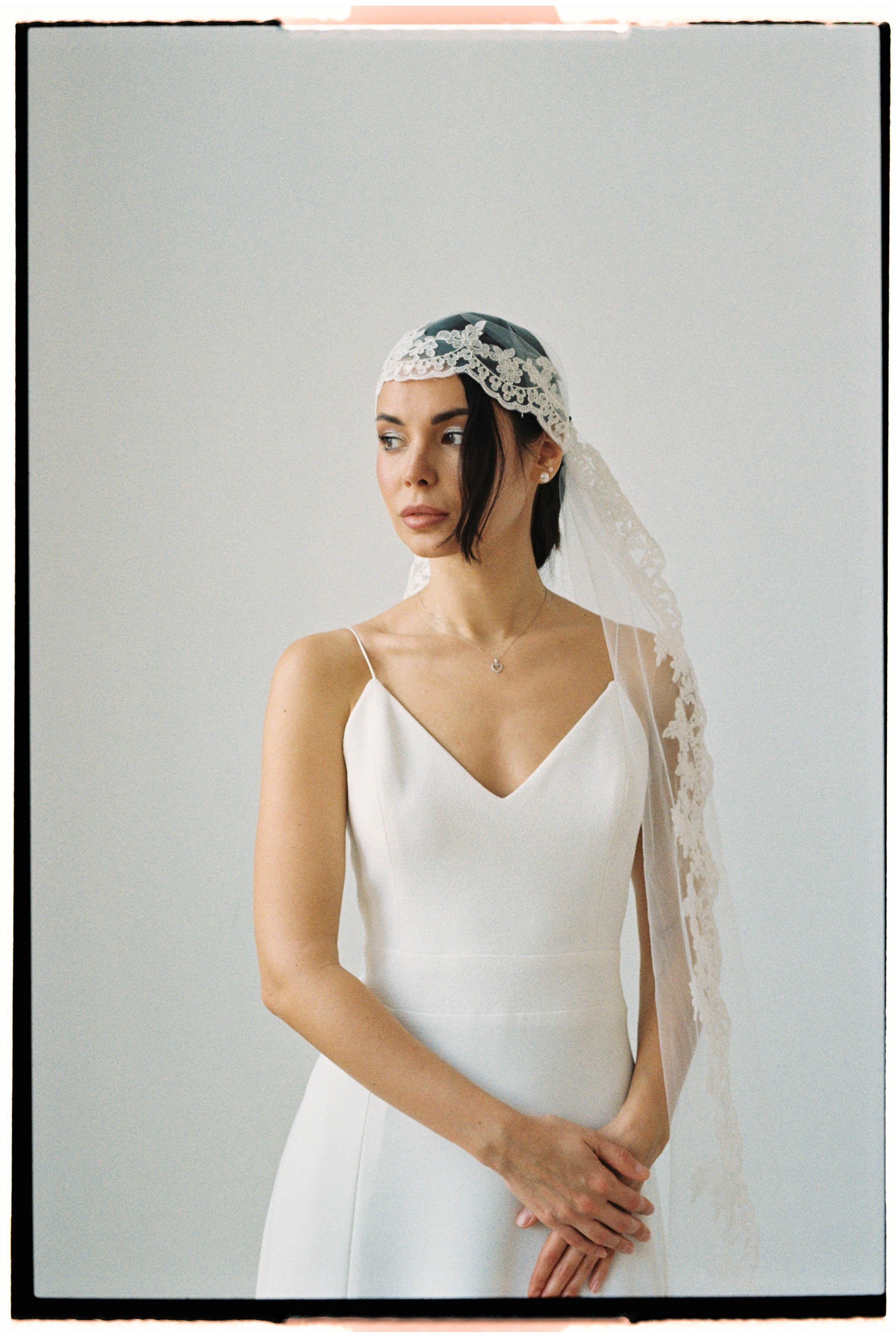 Mujer con vestido blanco de novia, usando un velo de encaje y pendientes pequeños, posando con las manos juntas, en un fondo gris claro.