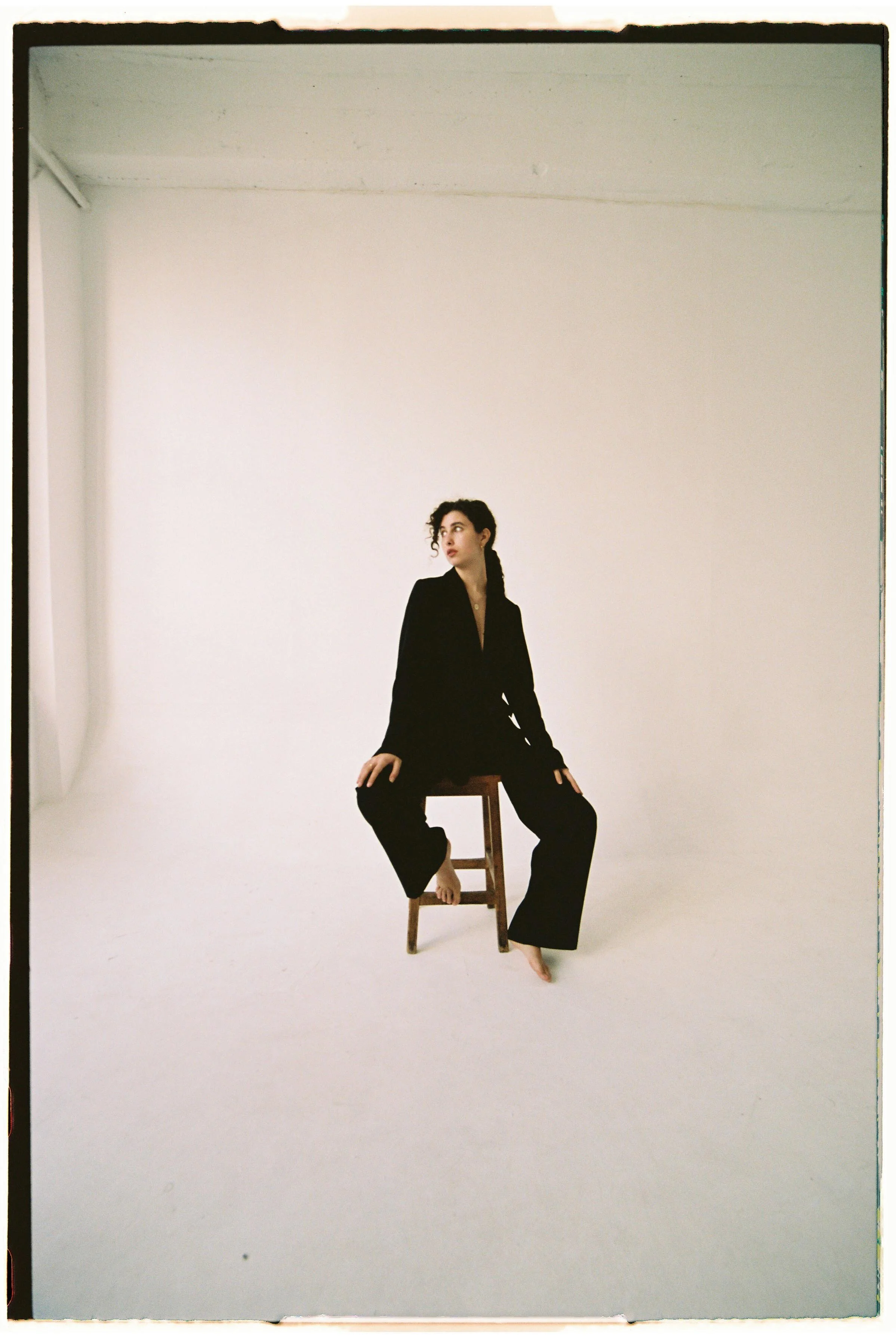 Una mujer con traje negro sentado en una silla de madera en un espacio amplio y vacío, con paredes blancas y sin otros objetos visibles.