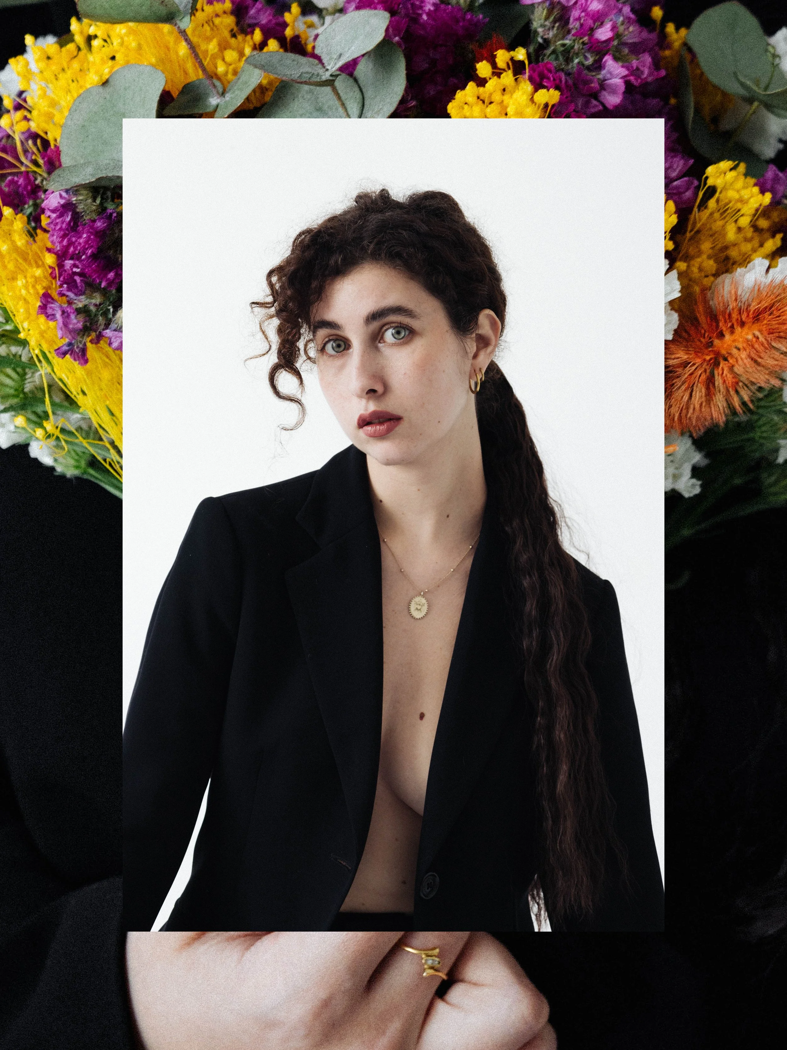 Retrato de una mujer con cabello rizado, ojos claros y piel clara, vistiendo un saco negro y joyería dorada, sobre un fondo blanco, con un marco de flores coloridas en el entorno.
