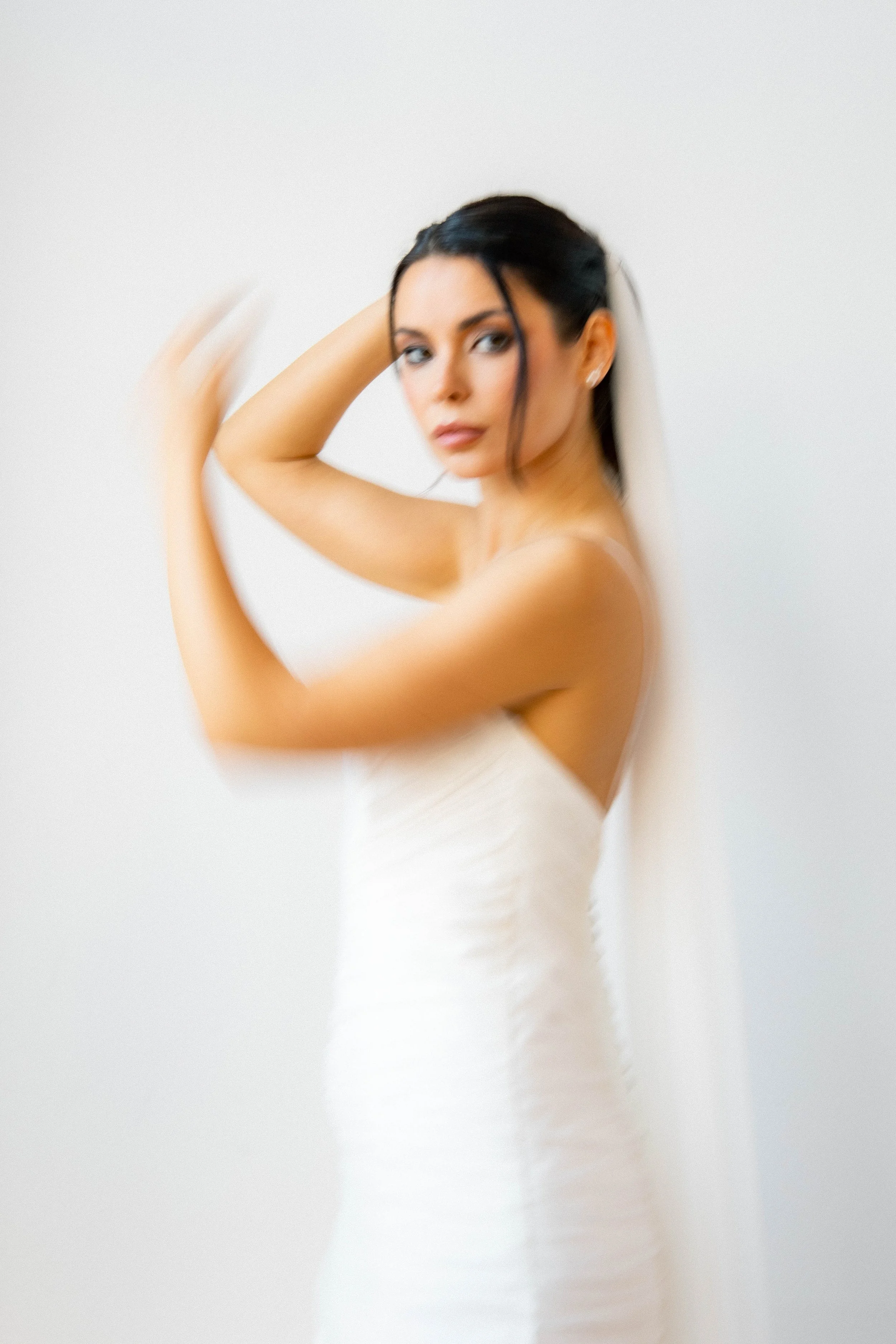 Mujer con vestido blanco y velo, en posición de posar, con cabello oscuro y expresión seria, contra fondo blanco.