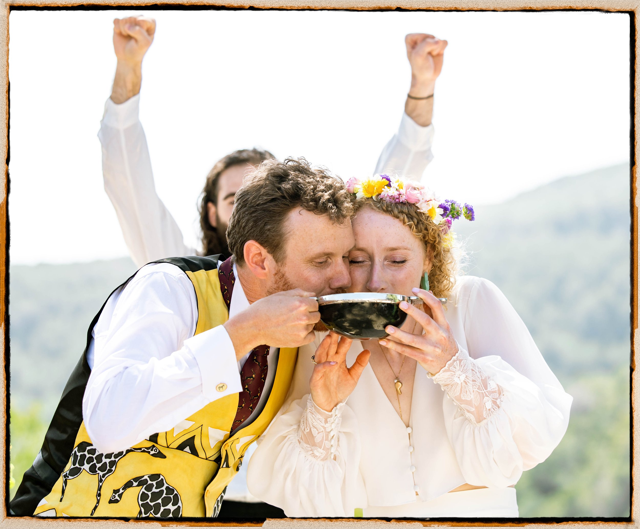 Una pareja de recién casados se besa y comparte una taza en una ceremonia al aire libre con un fondo de cerros y cielo claro, ella lleva una corona de flores y él un vestido formal.
