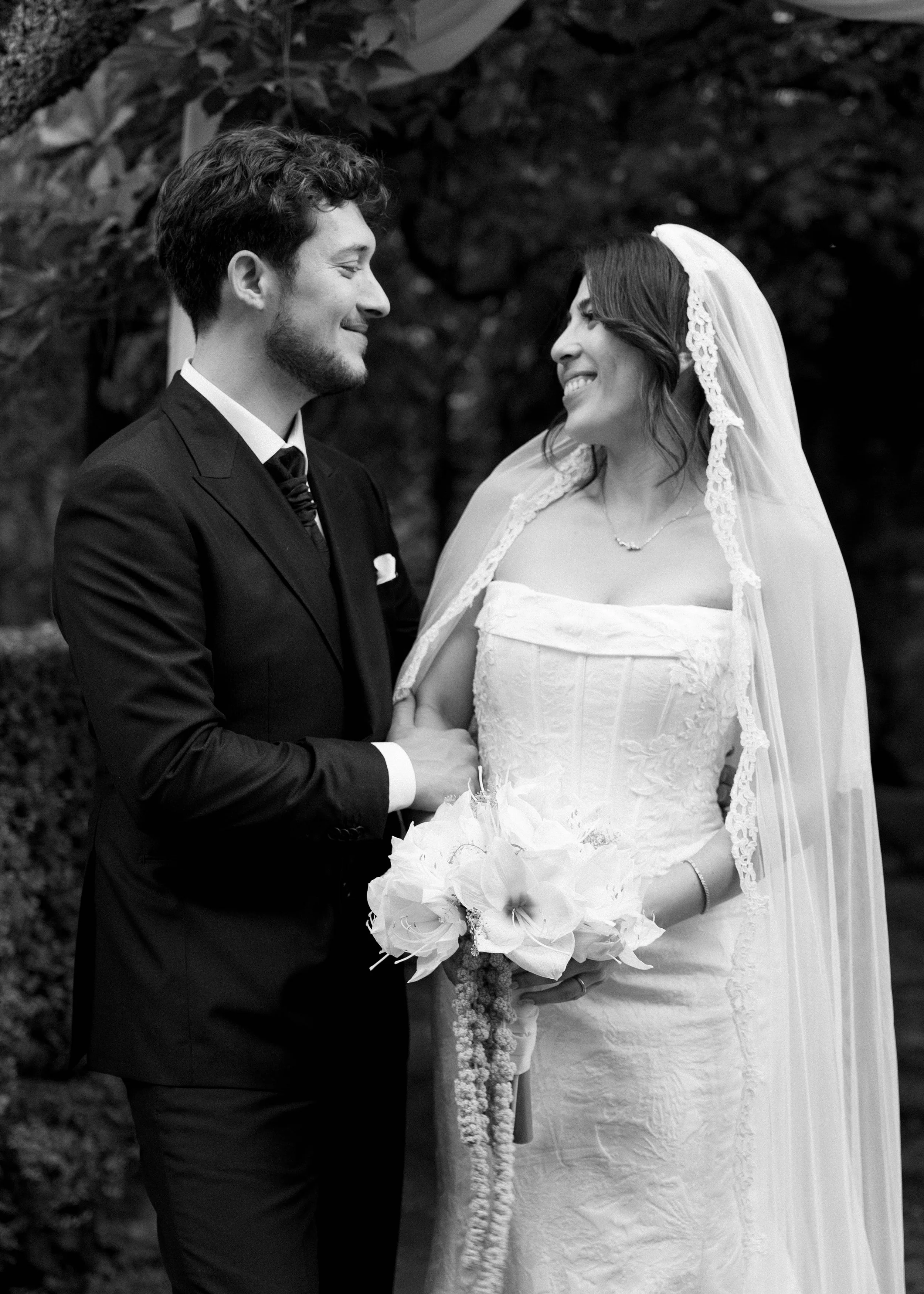 Pareja en boda, el hombre viste un esmoquin y la mujer un vestido de novia con velo, ambos sonriendo y mirándose, en un entorno natural.